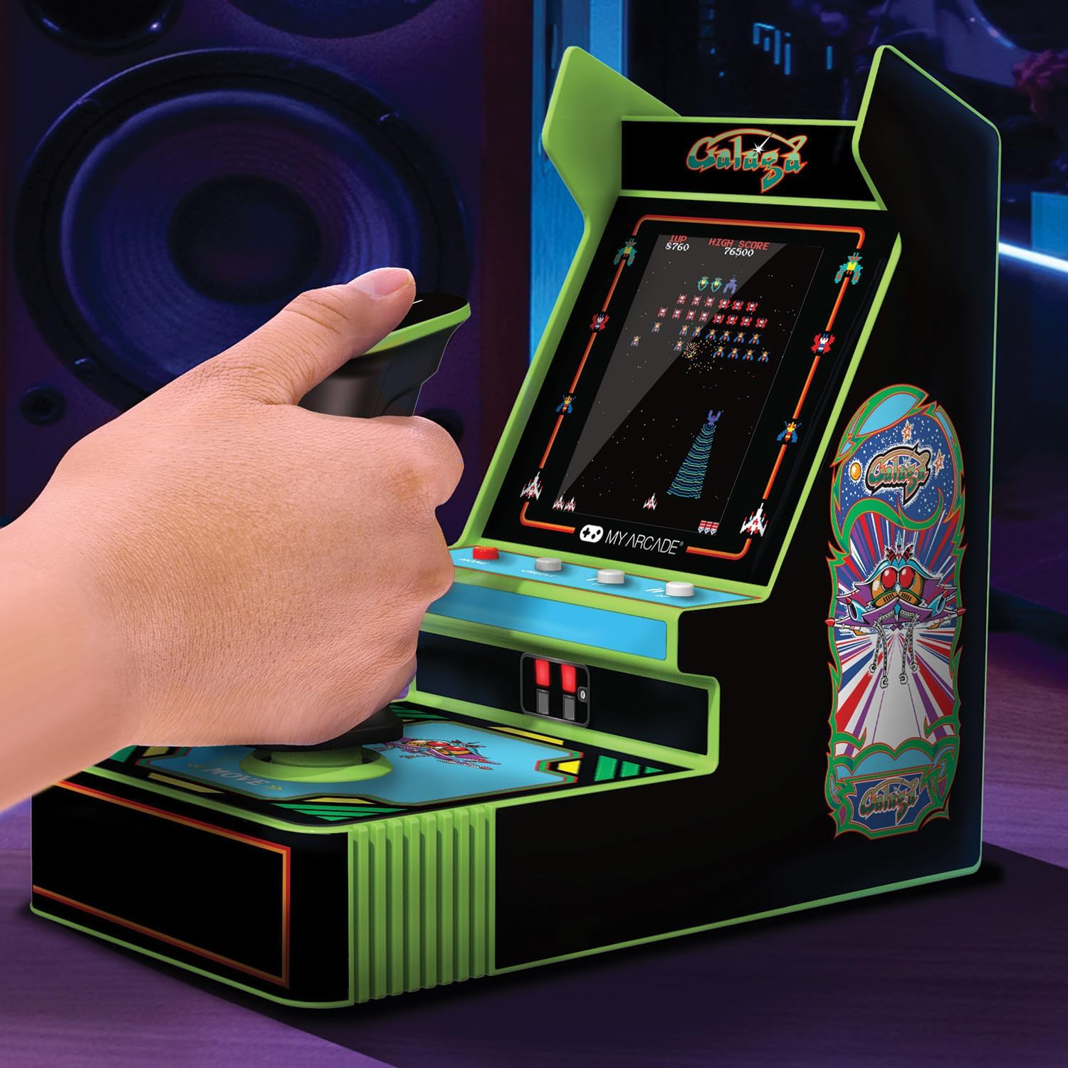 Una mano sostiene el joystick de un juego de arcade en miniatura. La pantalla muestra un juego de disparos espaciales.