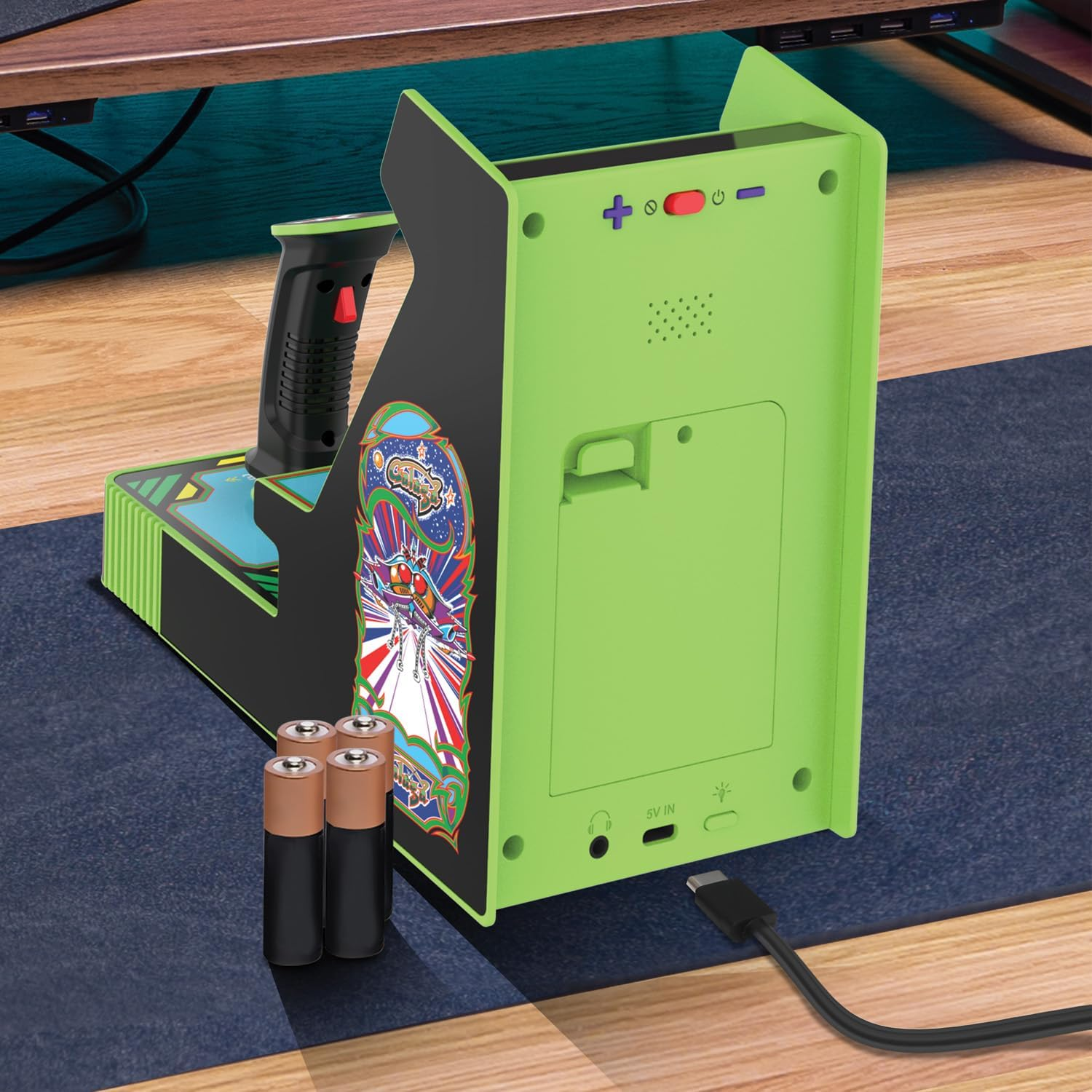 Un mini juego arcade verde y negro con joystick y baterías. Un cable USB está enchufado. Está sobre una alfombra azul.
