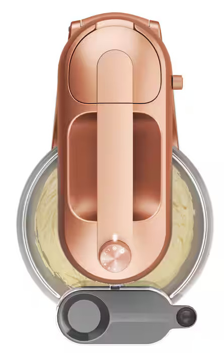 Miedziany mikser stojący, widok z góry. Szklana miska z żółtym ciastem poniżej. Widoczny przycisk i elementy sterujące.