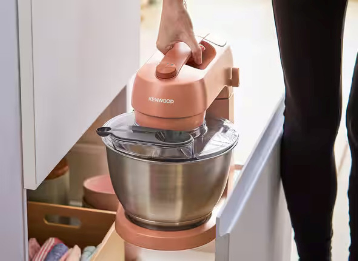 Osoba używa różowego miksera Kenwood na blacie kuchennym. Mikser ma misę ze stali nierdzewnej, przezroczystą plastikową pokrywę i jest różowy.