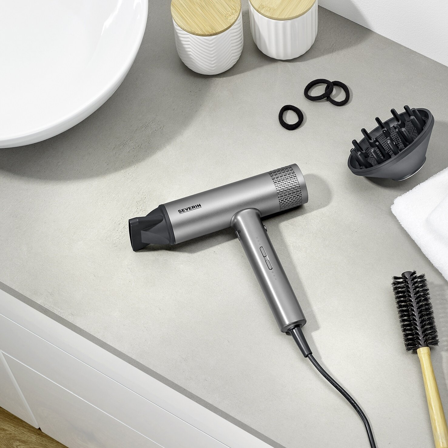 Szara suszarka do włosów, szczotka do włosów, gumki do włosów i dyfuzor leżą na szarym blacie. Biała umywalka i pojemniki w tle.