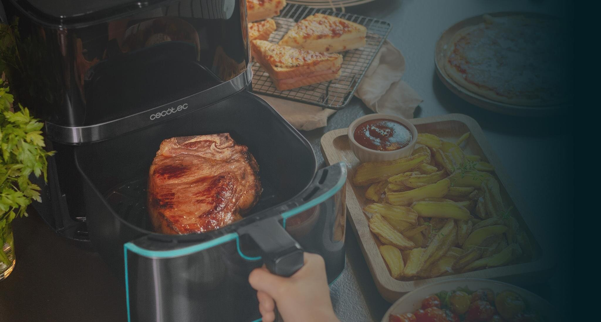 Frytkownica powietrzna z usmażonym stekiem. Inne dania to frytki, chleb i miska sosu. Ręka trzyma frytkownicę.