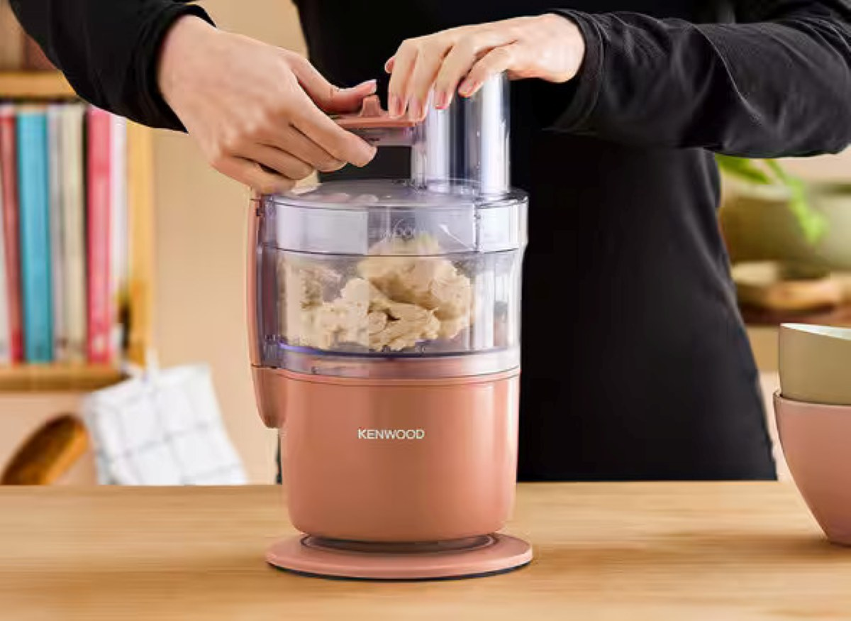 Osoba obsługuje różowy robot kuchenny Kenwood. Miska zawiera jedzenie. Regały na książki i miski w tle.