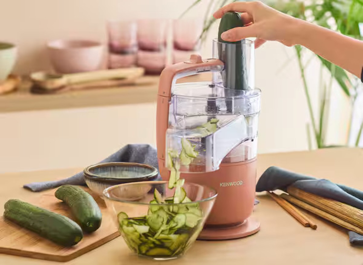 Osoba kroi ogórka za pomocą robota kuchennego Kenwood. Pokrojone ogórki wpadają do szklanej miski. Więcej ogórków leży na stole.