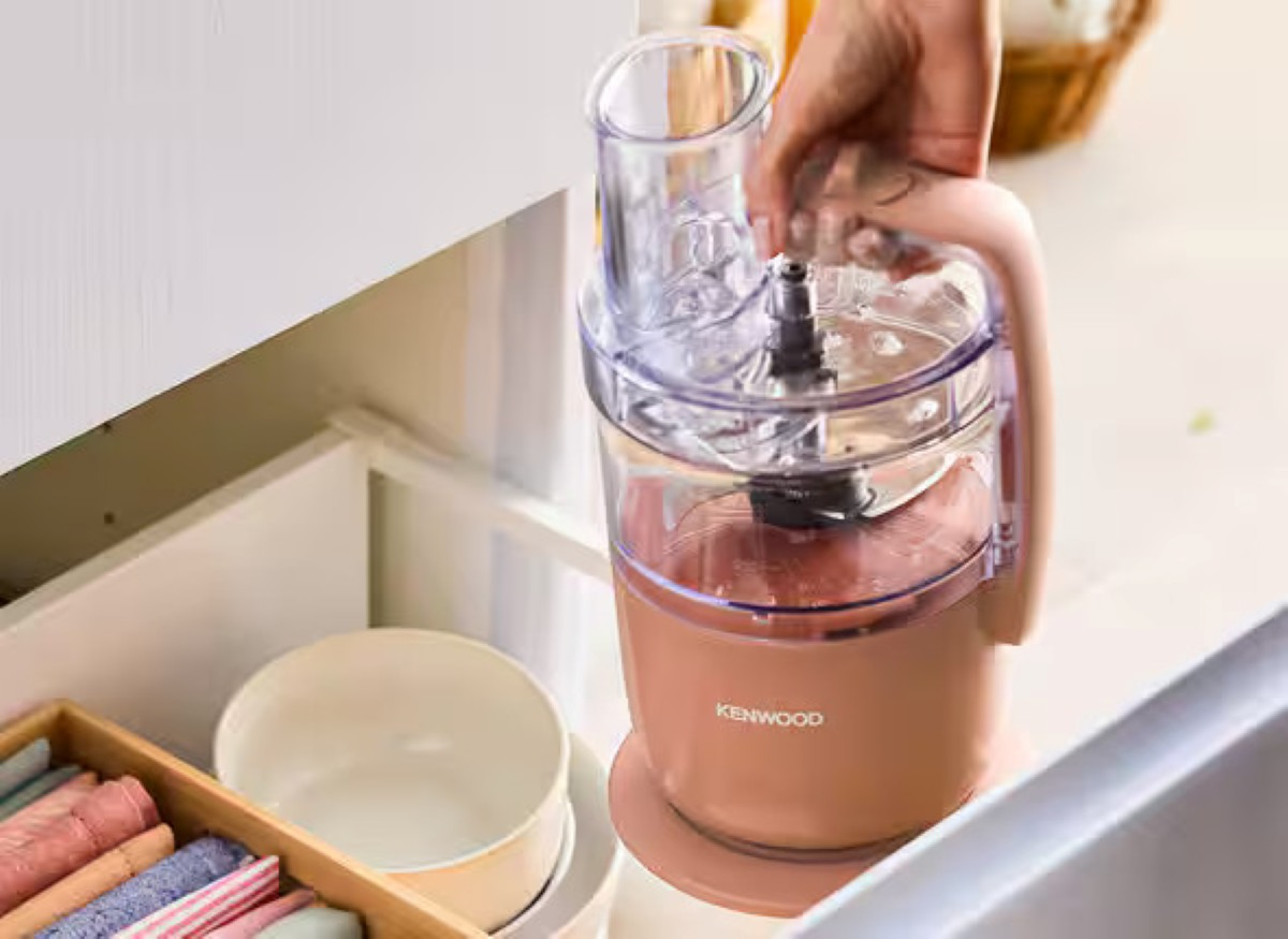 Robot kuchenny Kenwood trzymany jest przez rękę. Ma jasnoróżowy kolor, stoi w szufladzie, z innymi przedmiotami kuchennymi.