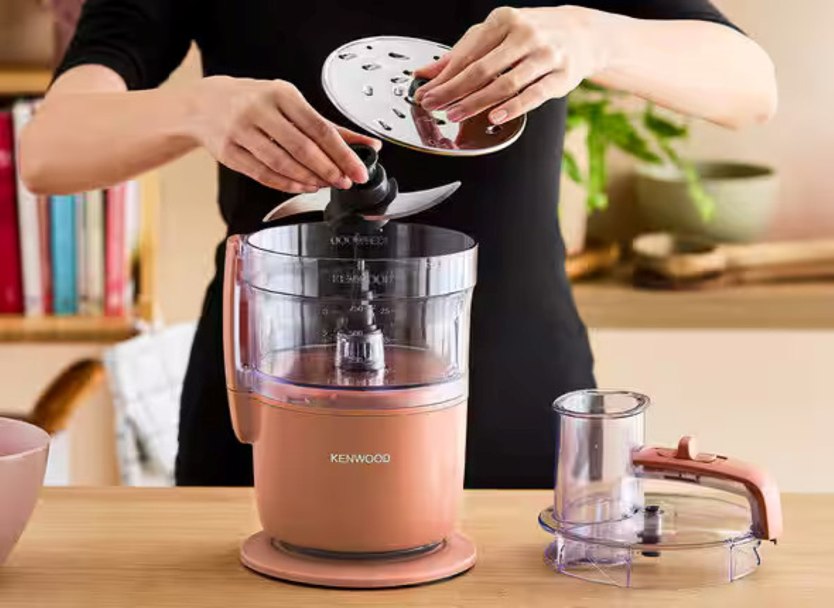 Osoba montuje robota kuchennego Kenwood. Ostrze, pokrywa i podstawa robota są widoczne na drewnianej powierzchni.