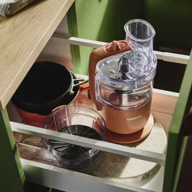 Szuflada kuchenna z robotem kuchennym, garnkiem i miską. Robot jest w kolorze brzoskwiniowym, garnek czarny, a miska przezroczysta.