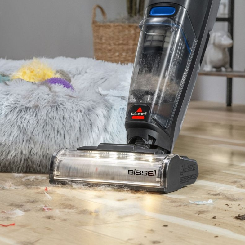 Odkurzacz Bissell czyści drewnianą podłogę z sierścią zwierząt. Puszek do spania dla zwierząt znajduje się w tle.