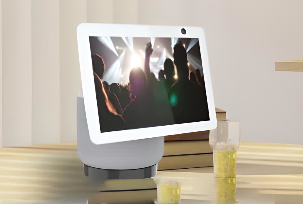 Inteligentny wyświetlacz pokazuje scenę z koncertu. Szklanka piwa i małe kubki stoją na drewnianym stole. Widoczny jest również stos książek.