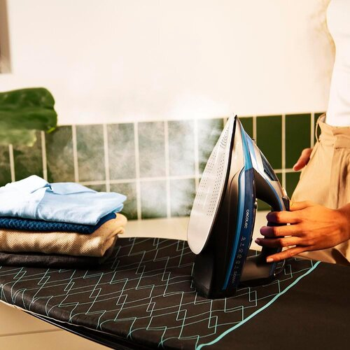 Osoba prasuje ubrania. Niebiesko-czarne żelazko emituje parę. Stos złożonych ubrań leży na desce do prasowania.