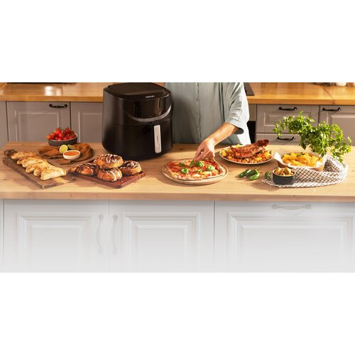 Na blacie kuchennym widnieje frytkownica powietrzna i różne potrawy: pizza, ciasta, frytki i warzywa. Osoba układa jedzenie.