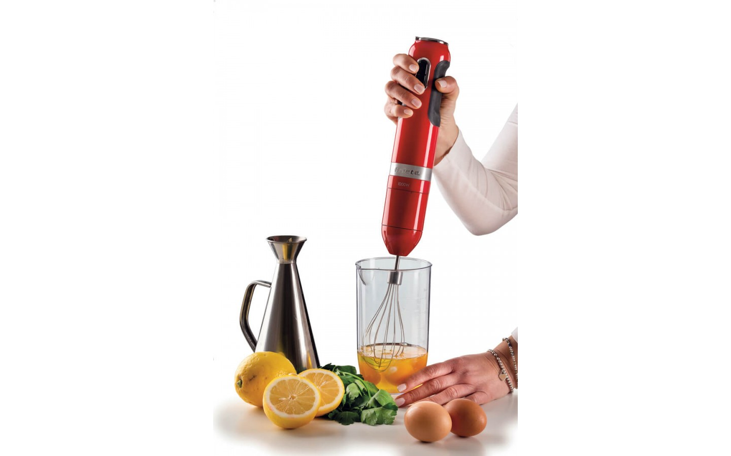 Ręka trzyma czerwony blender zanurzeniowy nad szklanką z trzepaczką. W pobliżu cytryny, jajka i oliwa z oliwek.