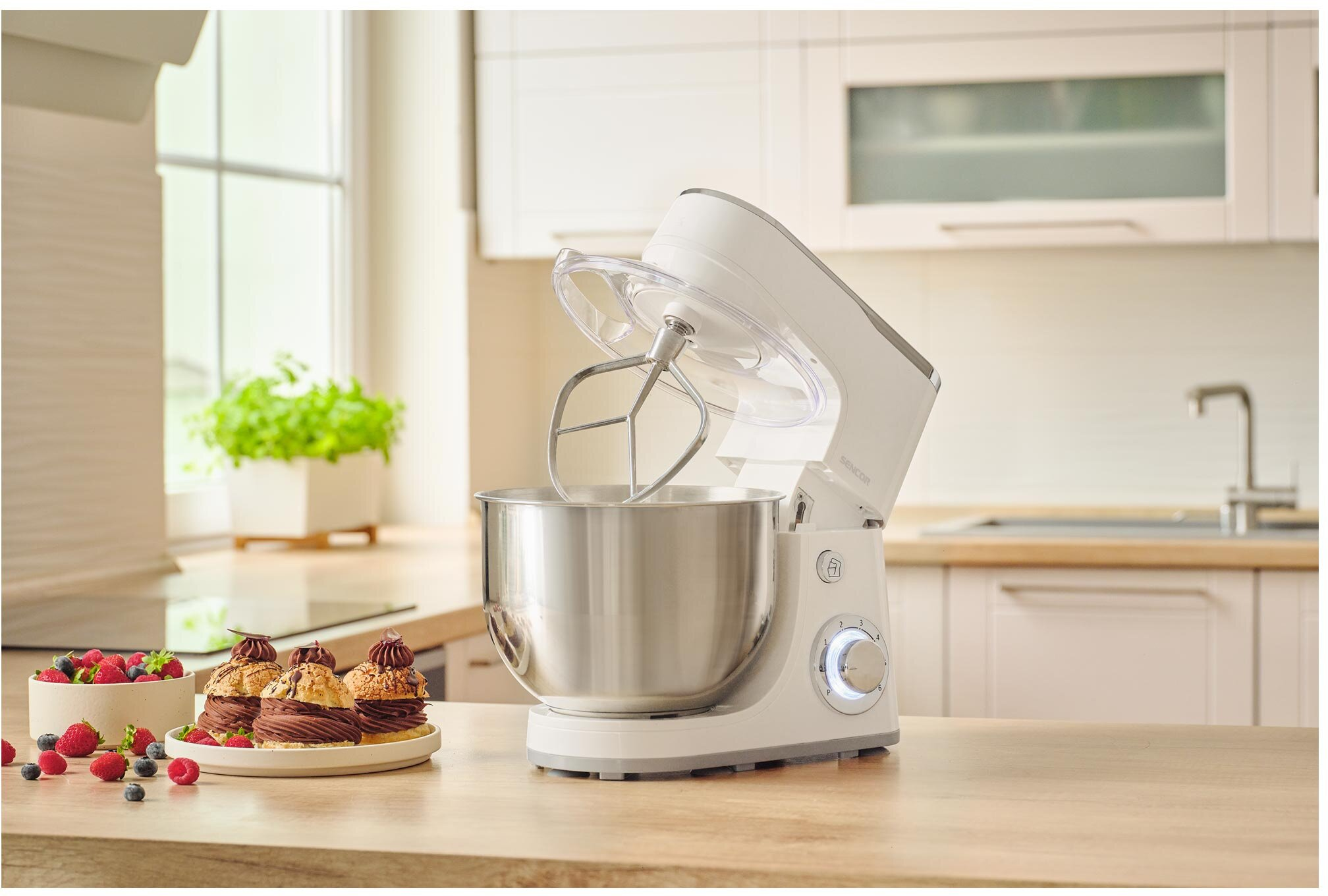 Biały mikser stojący z misą i ubijaczką stoi na drewnianym blacie. Ciasta i jagody przed mikserem.