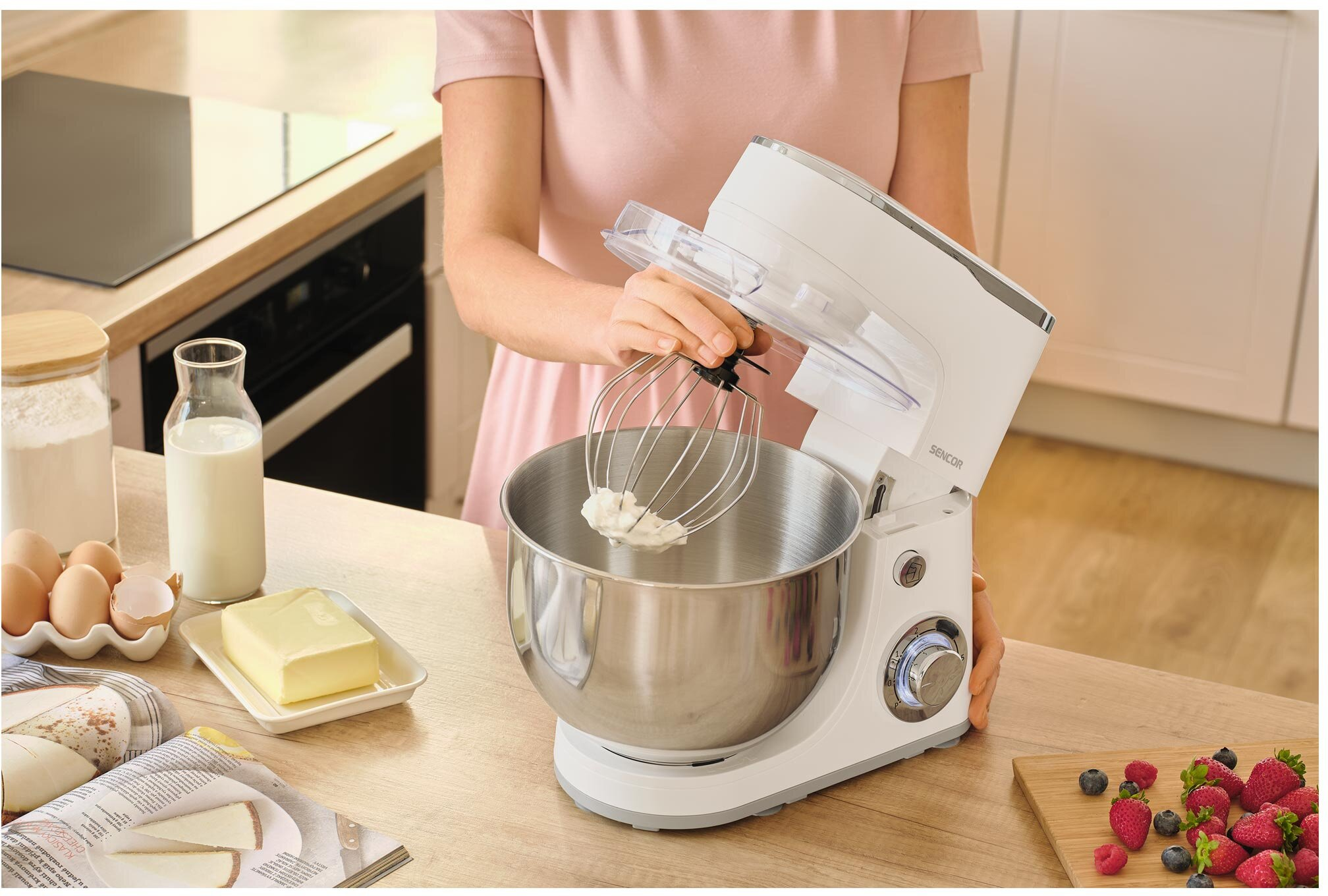 Osoba używa białego miksera stojącego ze trzepaczką. Na stole znajdują się składniki, takie jak jajka, mleko, masło i jagody.