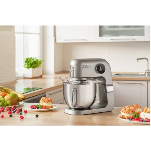 Srebrny mikser stojący na drewnianym blacie. Ciasta, owoce i jagody są wokół niego. Kuchnia z oknem w tle.