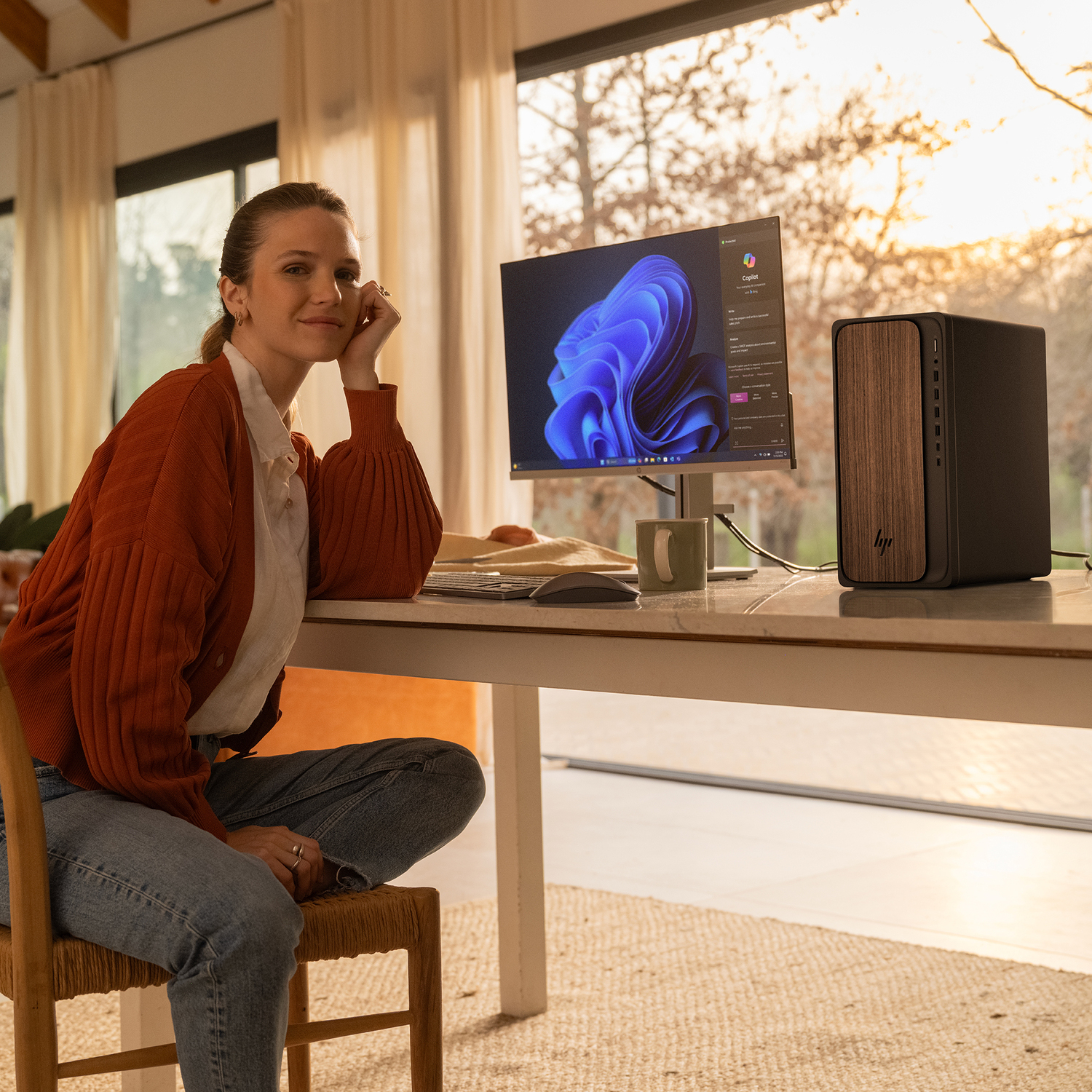 Mujer con cárdigan naranja sentada cerca de una computadora. Un monitor muestra un remolino azul. Una PC con frente de madera y una taza están en el escritorio.