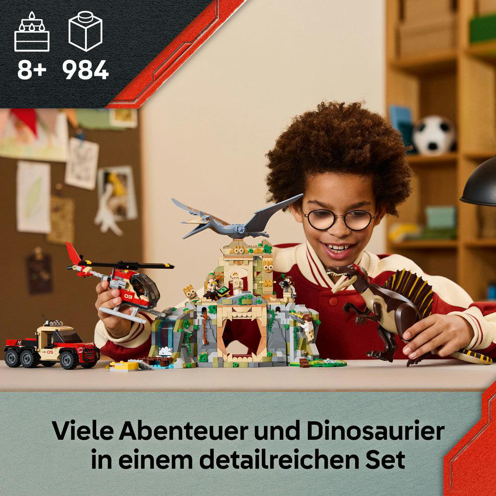 Ein Junge mit Brille hält ein Dinosaurierspielzeug und ein Modellgebäude. Links sind ein Hubschrauber und ein Lastwagen.