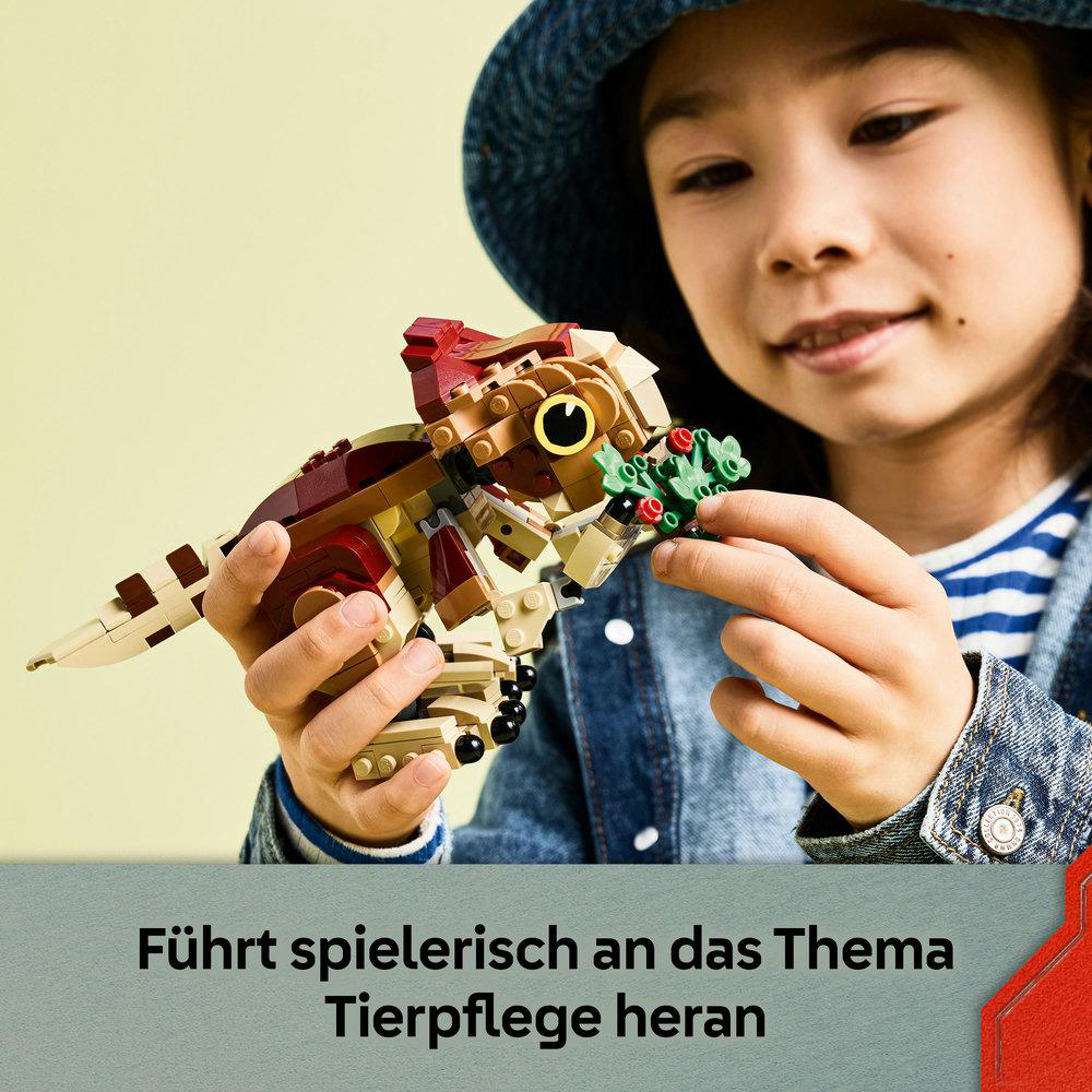 Ein Kind hält ein Lego-Dinosaurier-Spielzeug. Das Spielzeug ist braun, rot und hellbraun. Das Kind lächelt und trägt eine Jeansjacke und einen Hut.