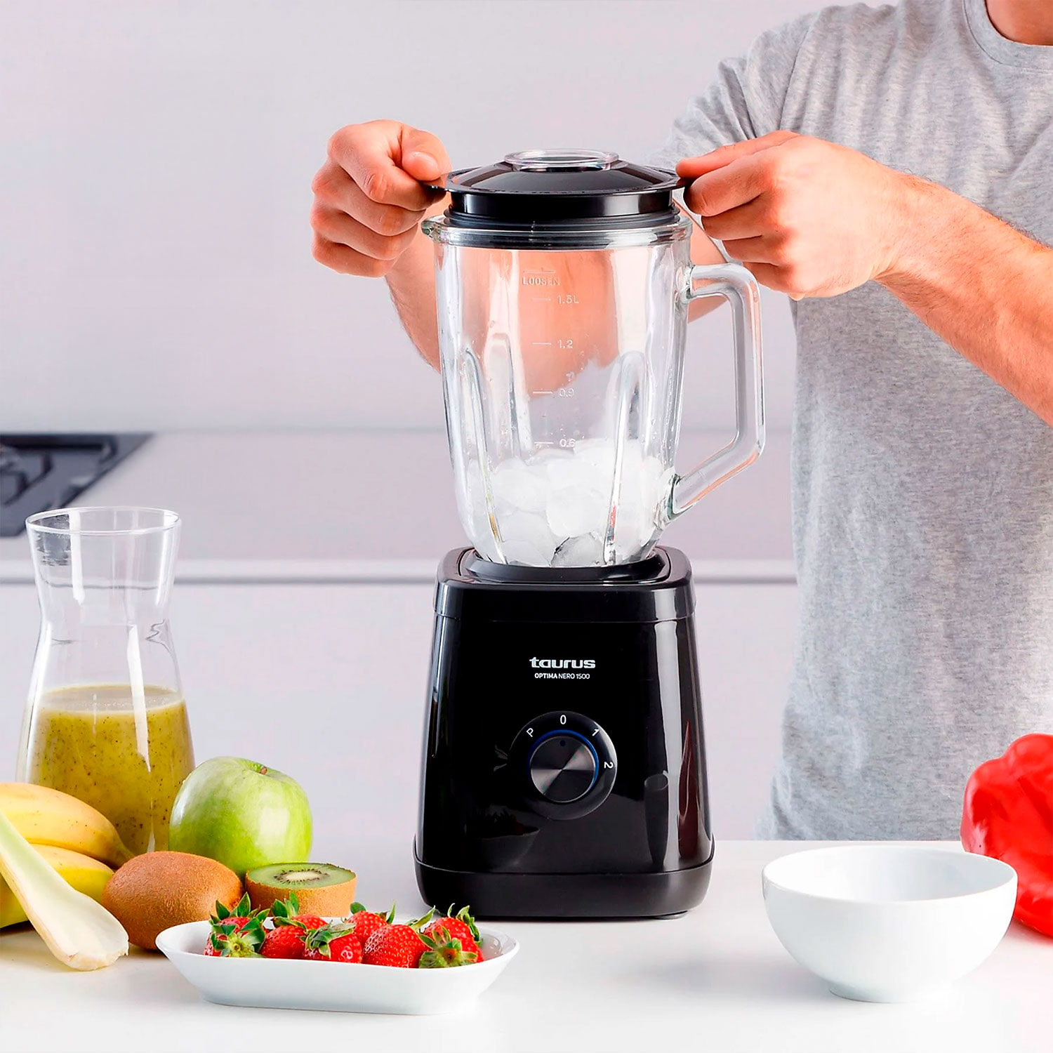 Una persona está preparando un batido. Una licuadora con hielo está sobre una mesa con frutas y verduras. Una persona está poniendo la tapa.