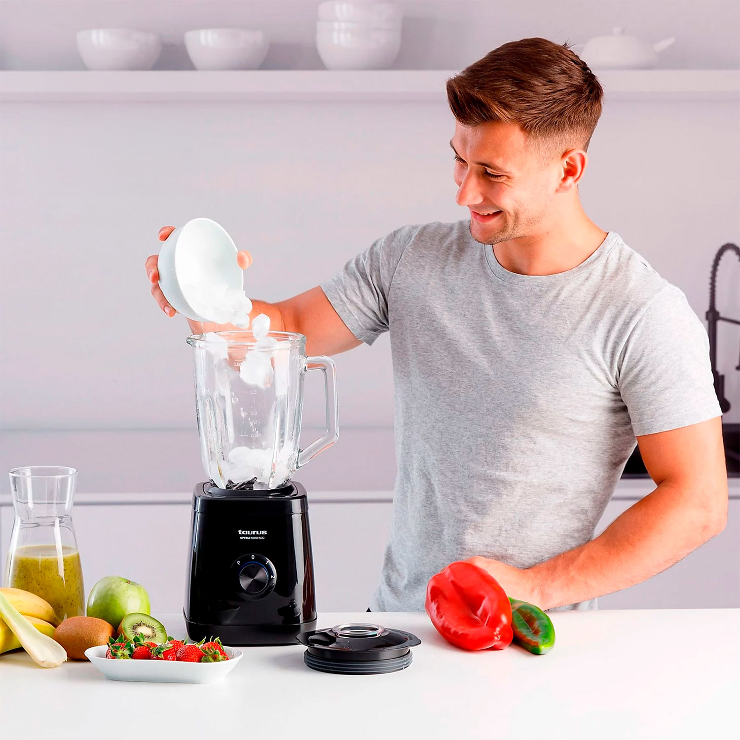 Un hombre vierte hielo de un cuenco en una licuadora. Frutas como fresas, kiwi, manzana y plátanos están en la encimera.