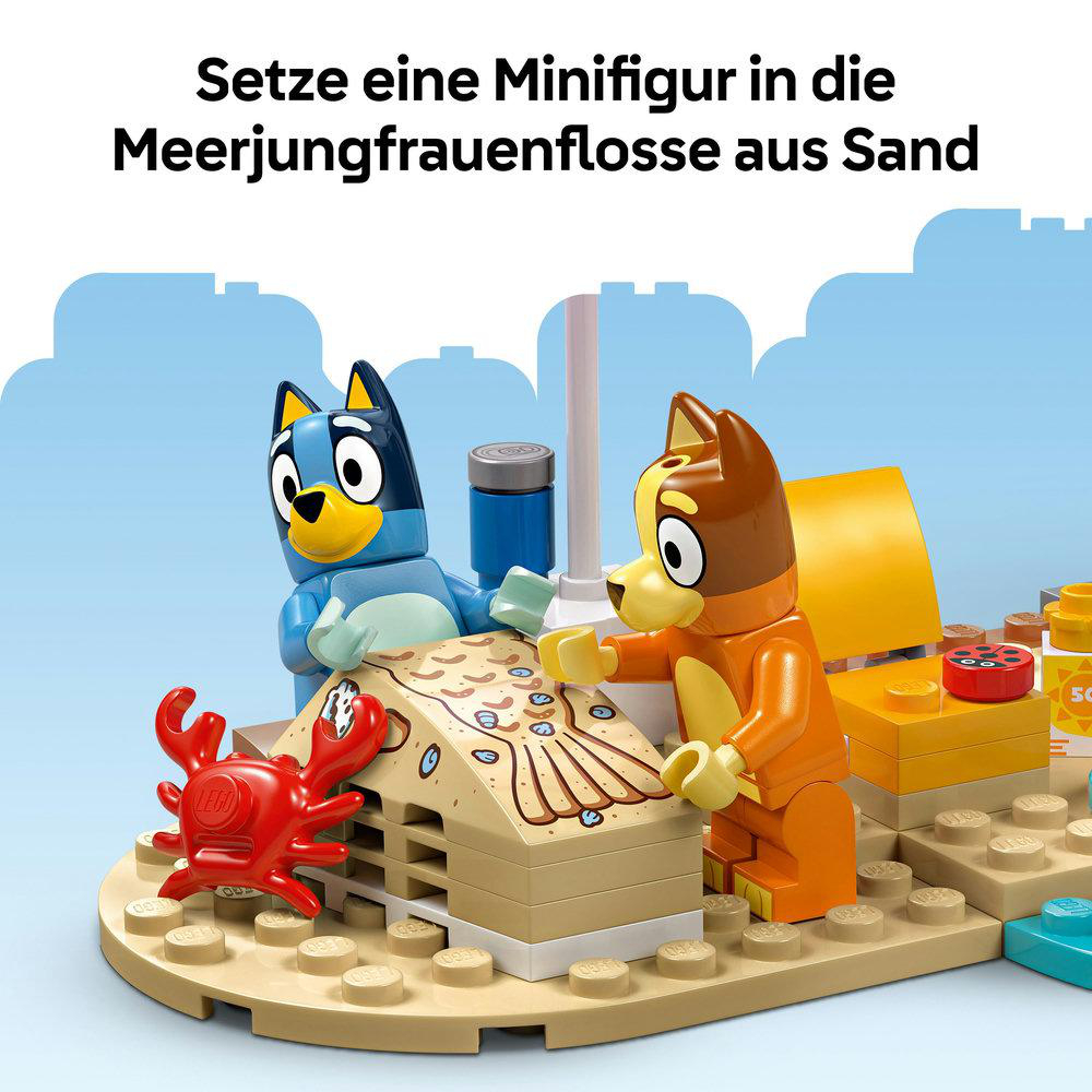 LEGO-Figuren von Bluey und Bingo am Strand mit einer Sand-Meerjungfrauenflosse und einer Krabbe. Ein blauer Kühler ist in der Nähe.