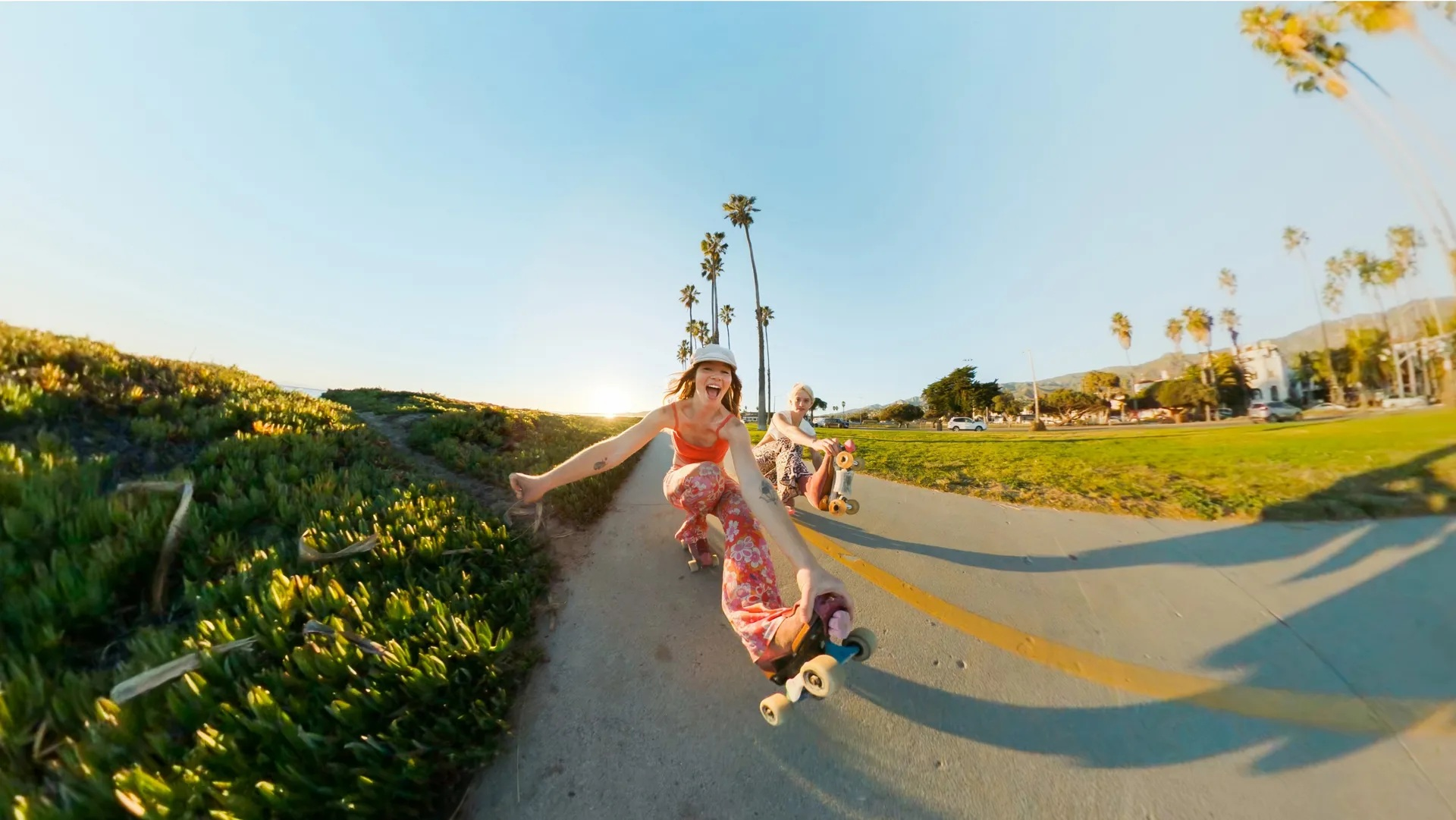 Dwie kobiety jeżdżą na deskorolce po ścieżce. Drzewa palmowe wzdłuż ścieżki. Jedna kobieta uśmiecha się do kamery. Zielona trawa i błękitne niebo.