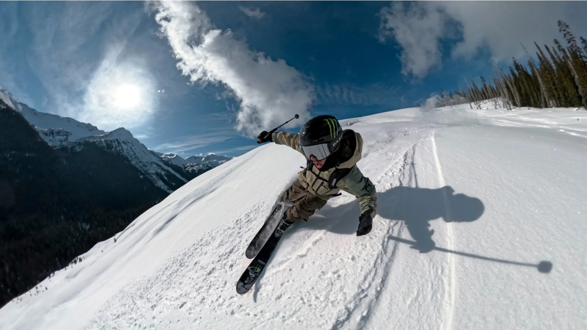 Narciarz zjeżdża po zaśnieżonym stoku w jasnym słońcu. W tle góry i drzewa. Narciarz ma na sobie kask i gogle.