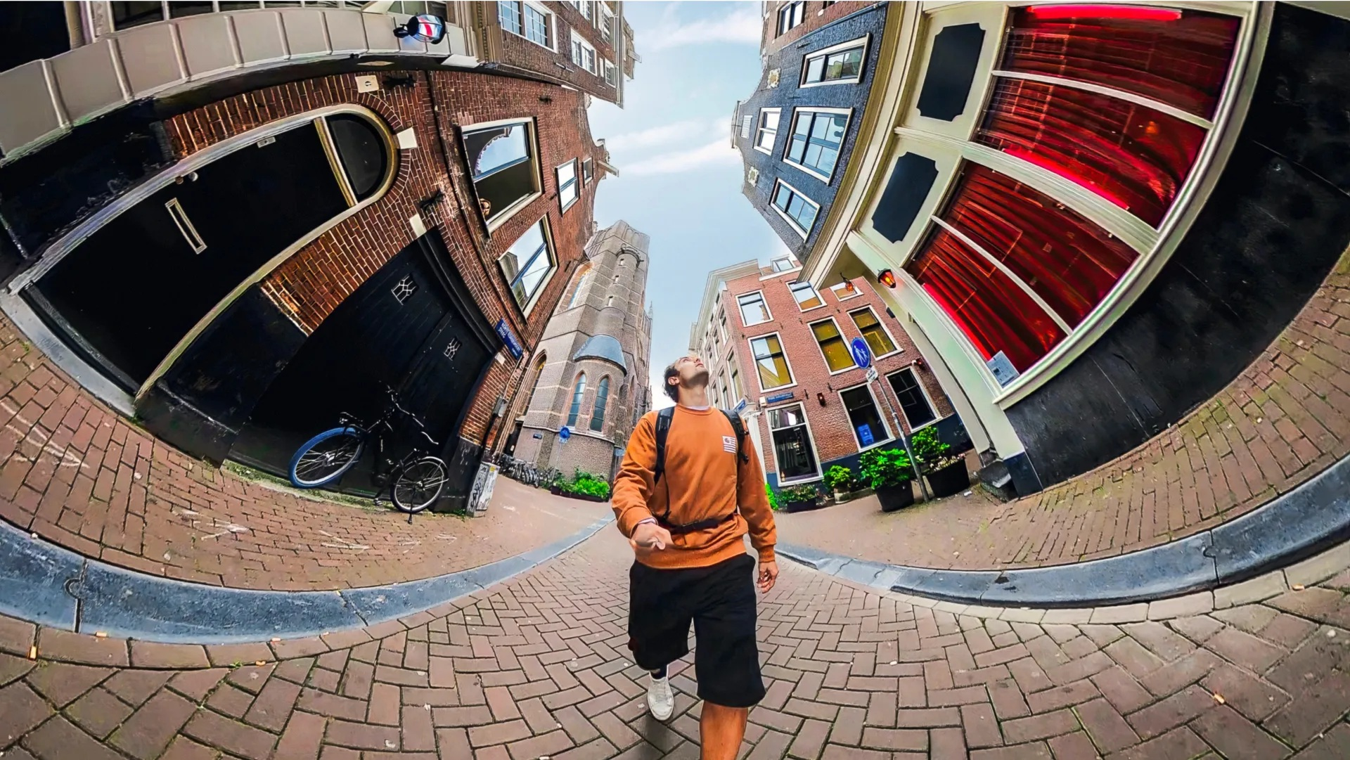 Mężczyzna idzie ceglaną ulicą, patrząc w górę. Budynki obramowują ulicę. Rower zaparkowany z boku. Niebo jest niebieskie.