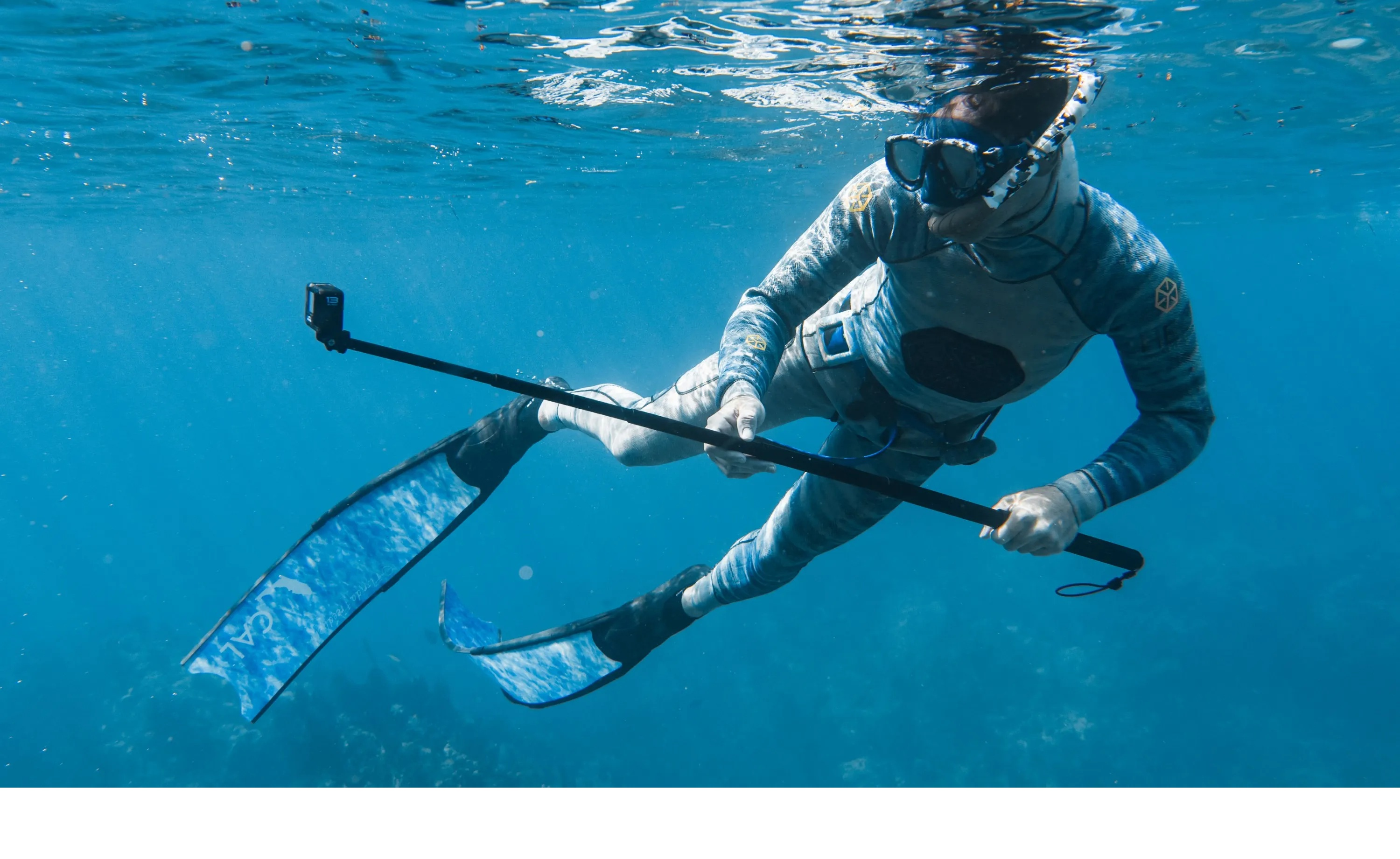 Osoba w kombinezonie do nurkowania pływa pod wodą z płetwami i kamerą na patyku. Woda jest niebieska, a światło słoneczne się filtruje.