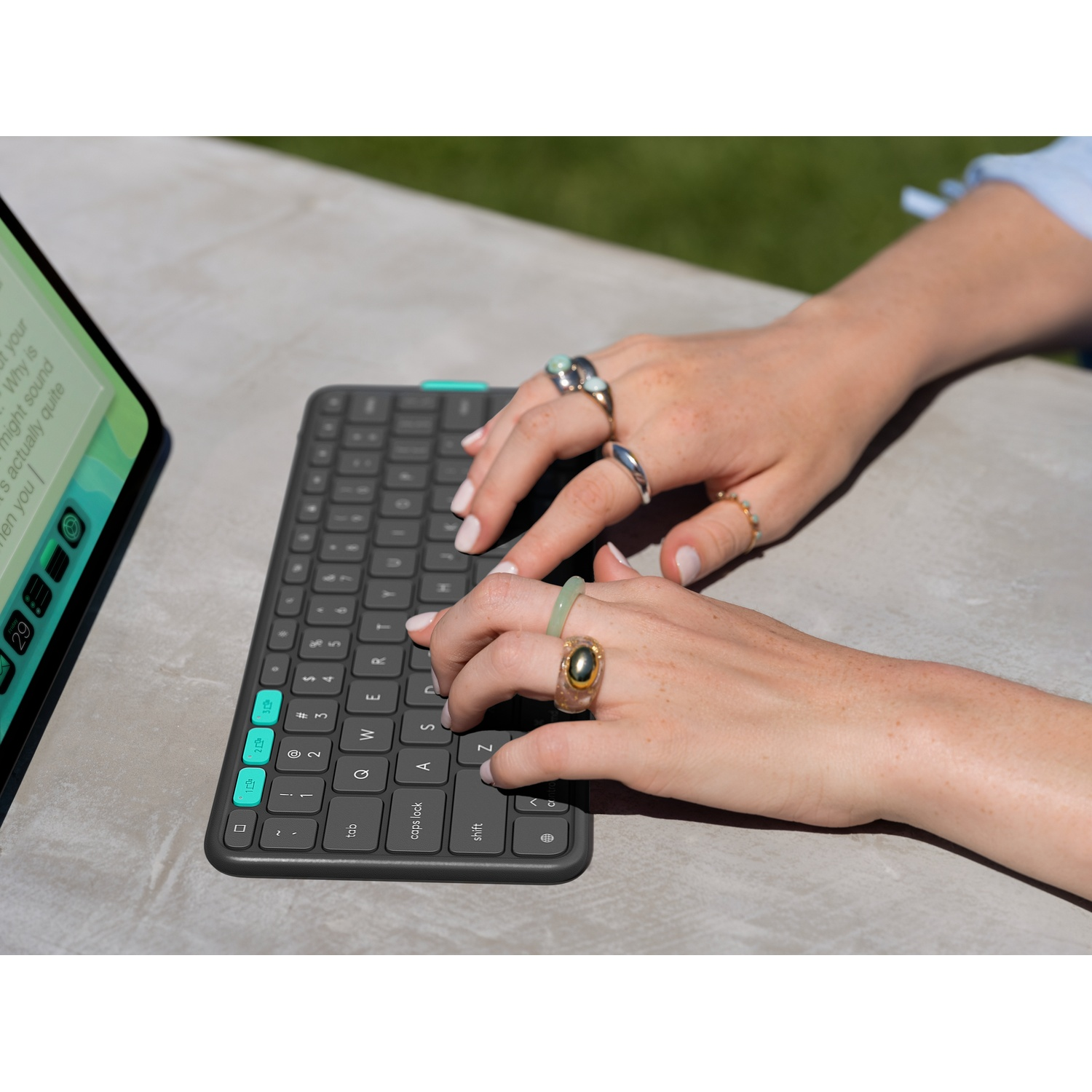 Mani con anelli che digitano su una tastiera grigia. Un tablet a sinistra. Lo sfondo è una superficie grigia e verde.