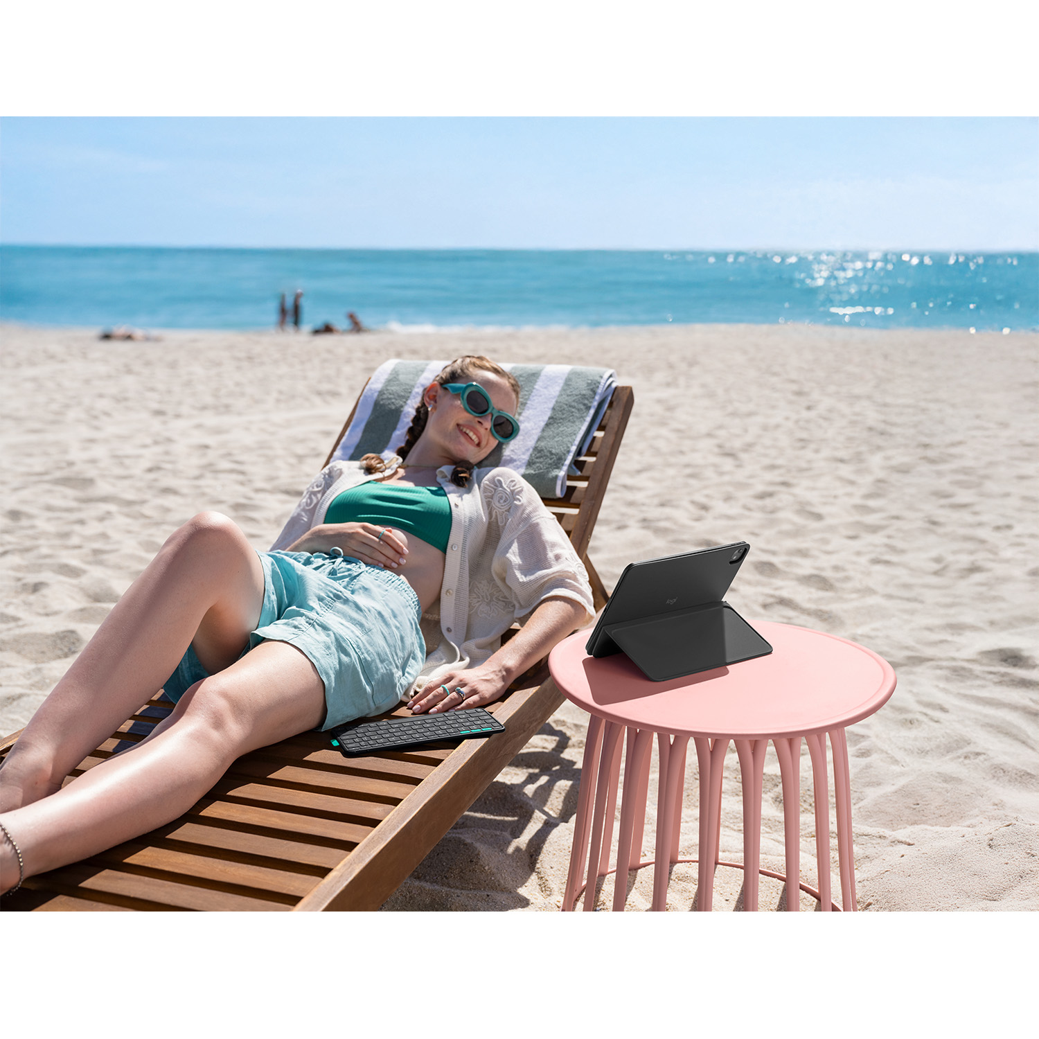 Donna sulla sedia a sdraio, occhiali da sole, pantaloncini e top. C'è una tastiera, un tablet e un tavolo rosa. Il mare è sullo sfondo.