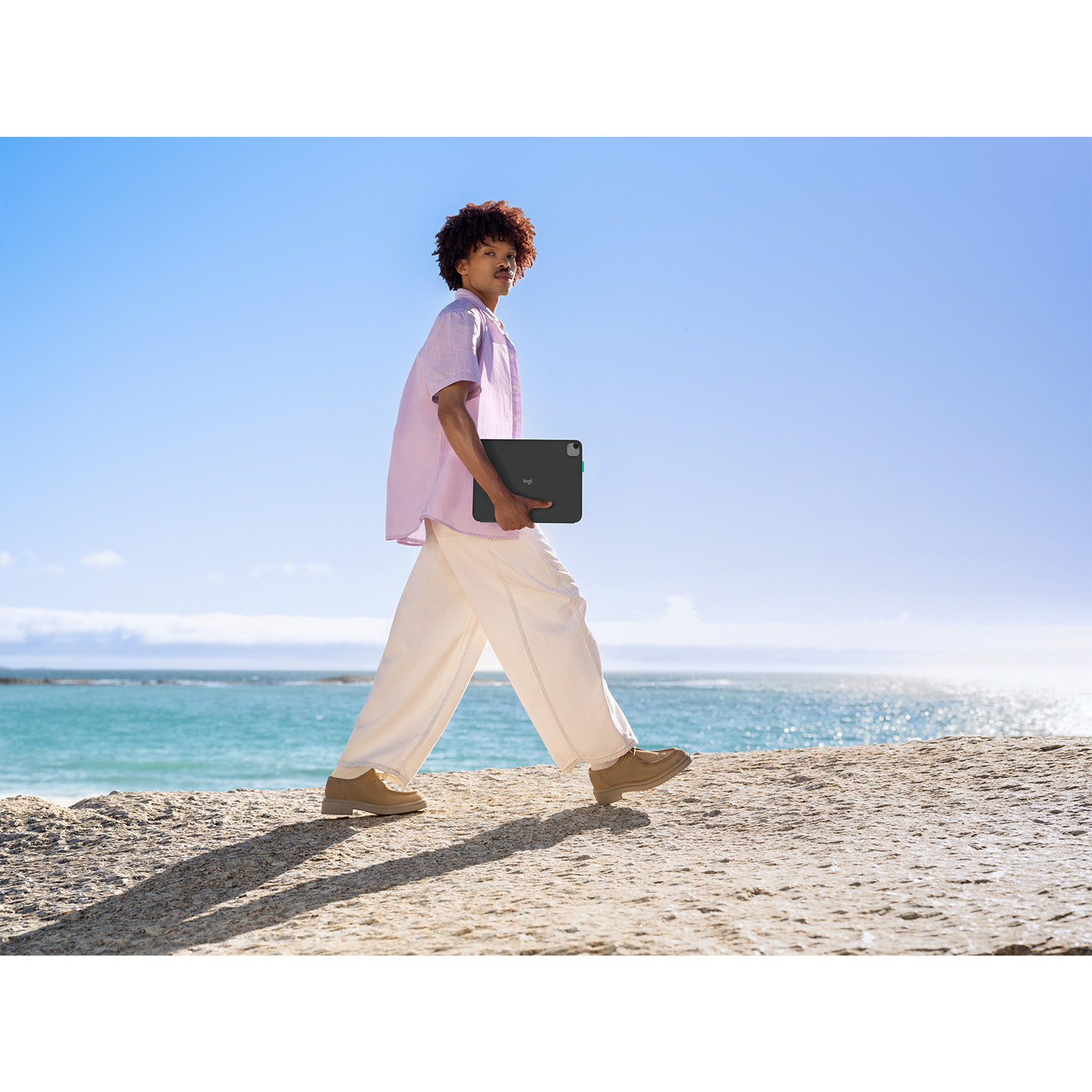 Una persona cammina sulla spiaggia tenendo in mano un tablet. Indossa una camicia rosa, pantaloni bianchi e scarpe marroni. Si vedono il mare e il cielo.