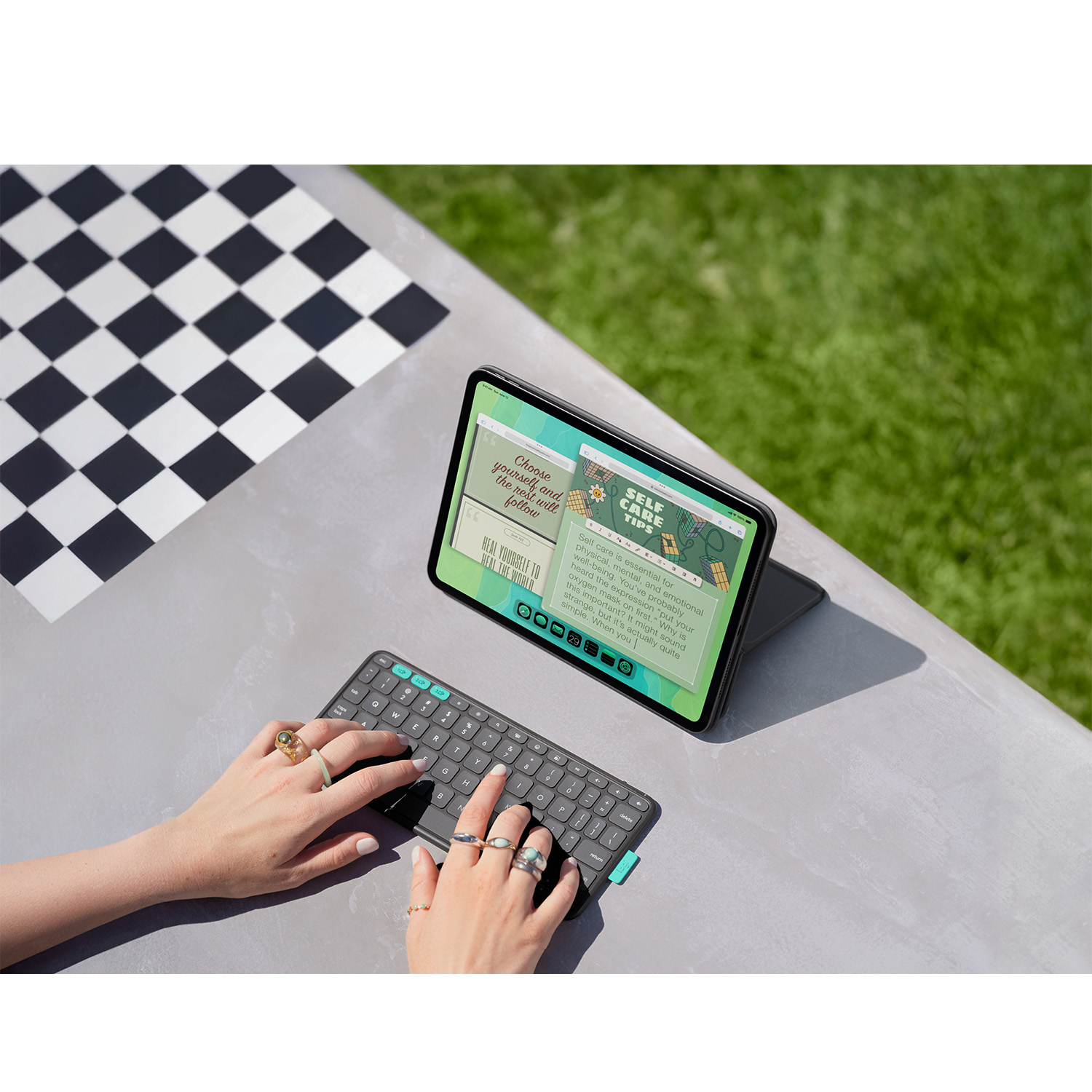 Mani che digitano su una tastiera con un iPad che mostra consigli per la cura di sé. Uno sfondo a scacchi ed erba verde sullo sfondo.