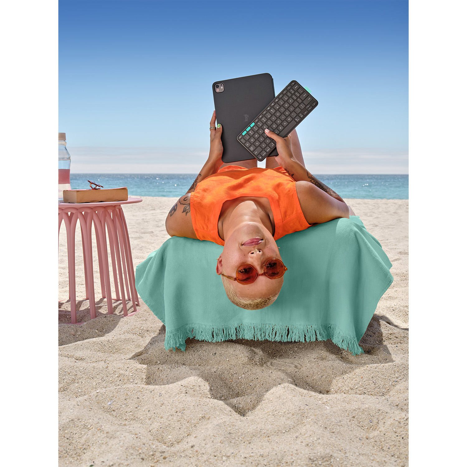 Persona sulla spiaggia con tablet e tastiera. Top arancione, tavolo rosa, mare e cielo blu. Relax su un asciugamano verde.