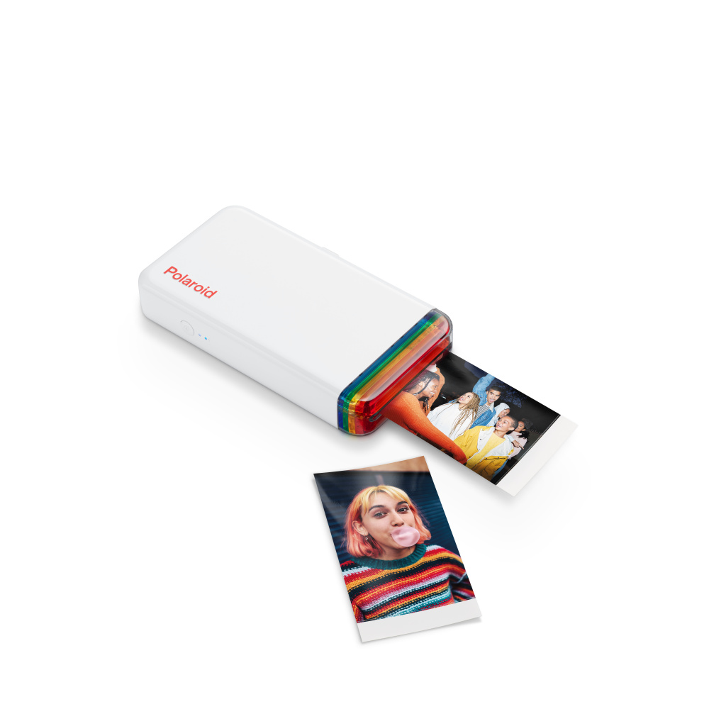 Una impresora Polaroid blanca con detalles de arcoíris imprime fotos. Se ven dos fotos: una de un grupo y otra de una mujer soplando una burbuja.