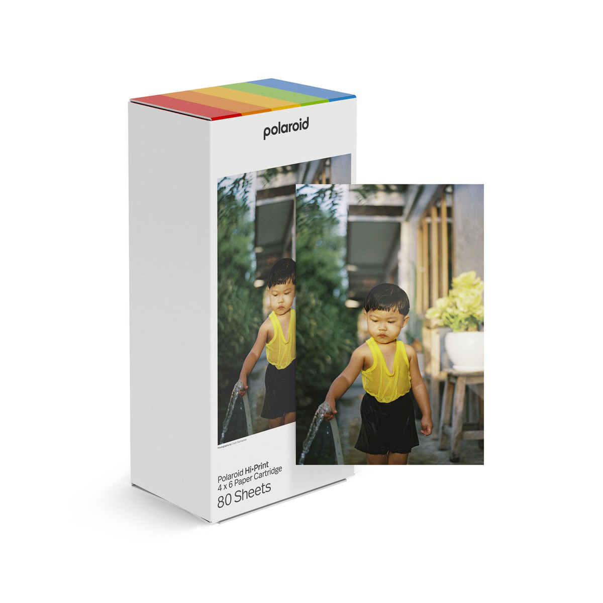 Un niño con camisa amarilla y pantalón corto negro sostiene una manguera, el agua rocía. Se muestra una caja Polaroid, con una imagen del niño.
