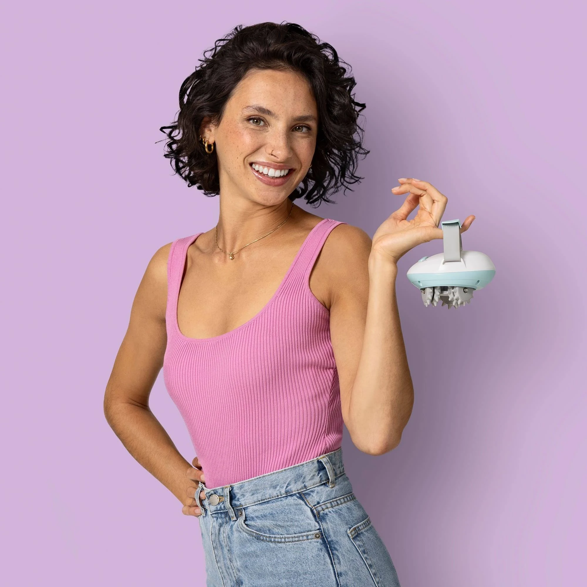 Mujer con top rosa sosteniendo un masajeador blanco y azul, sonriendo. Con jeans, sobre un fondo púrpura.