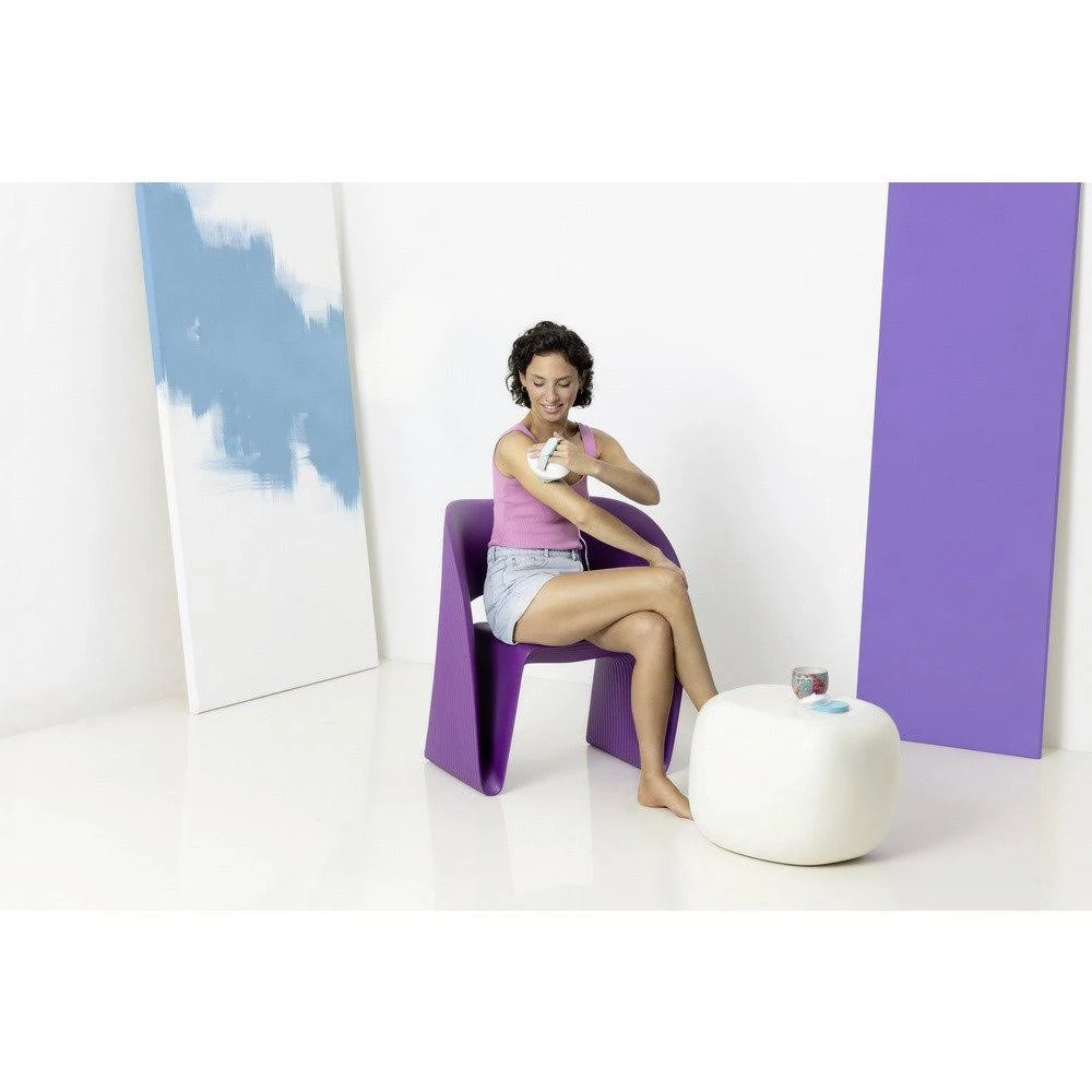 Una mujer sentada en una silla morada se aplica loción. Una mesa blanca con objetos está a su lado. El fondo incluye paredes blancas y paneles de color.