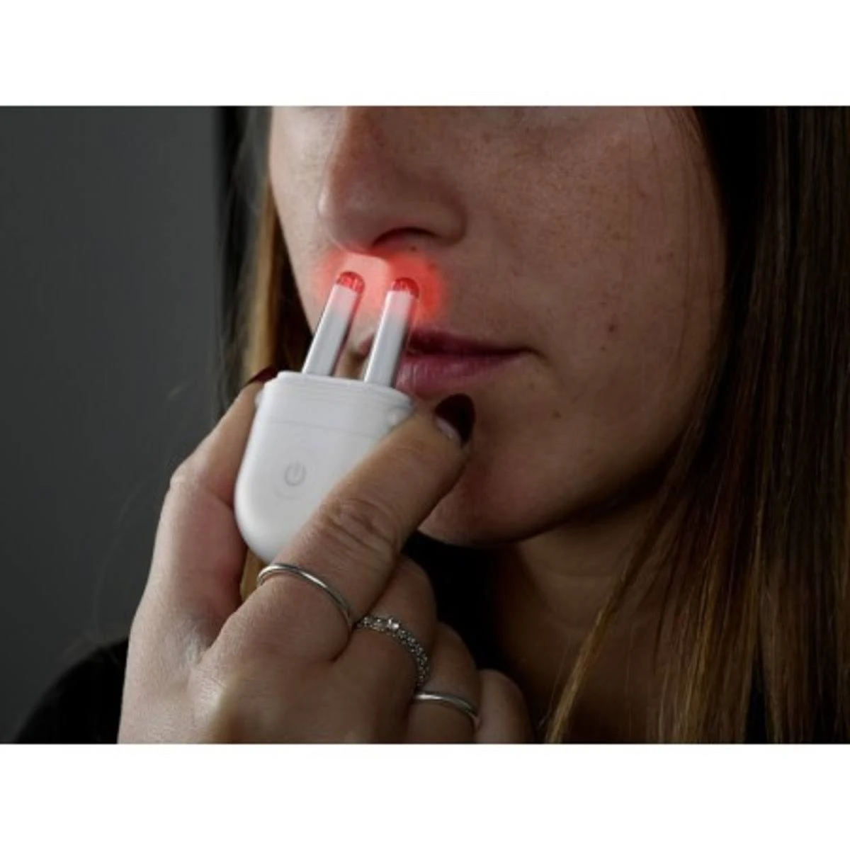 Una mujer sostiene un dispositivo blanco con puntas rojas brillantes cerca de su nariz. El dispositivo tiene un botón de encendido. Anillos adornan sus dedos.
