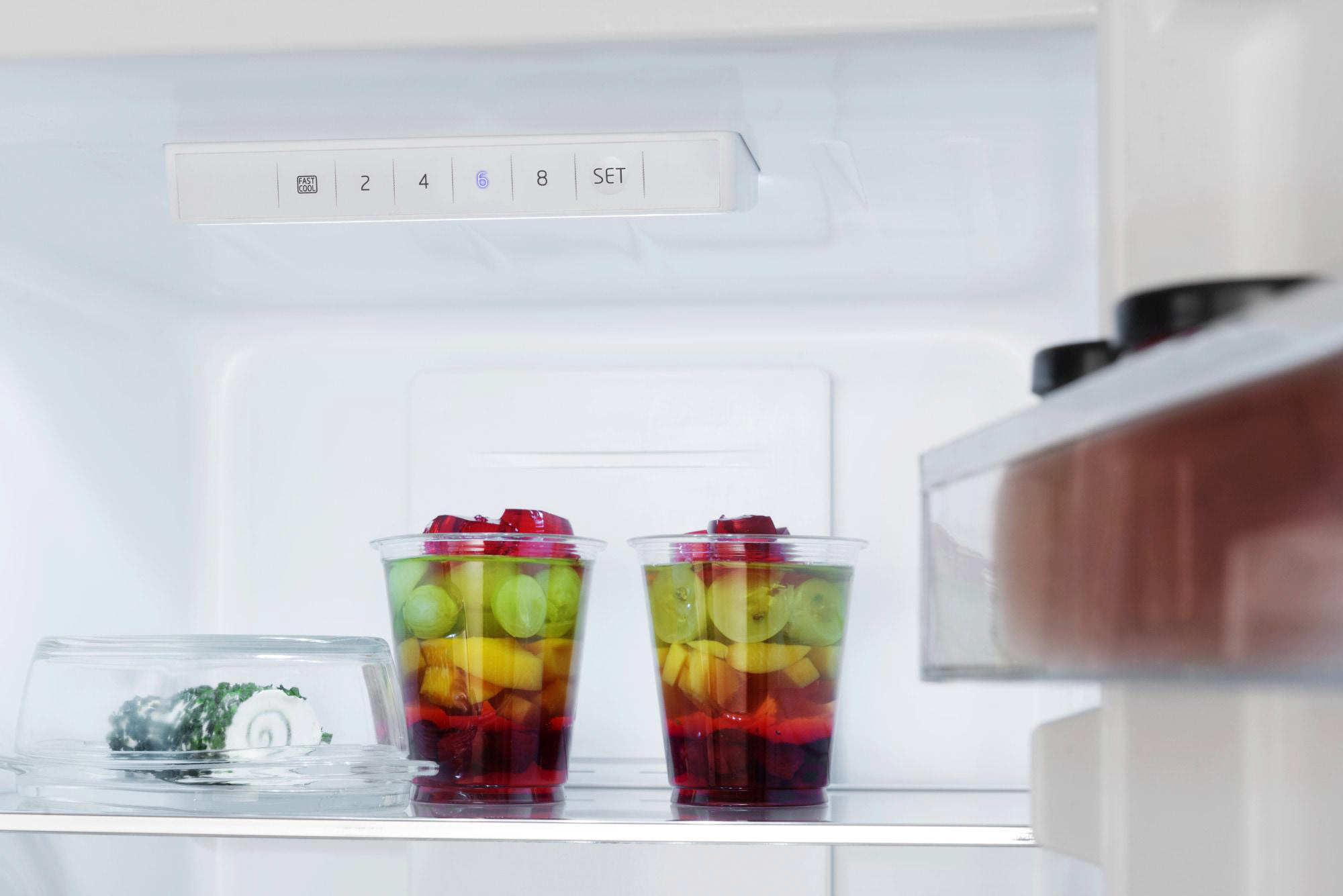 Im Kühlschrank sind zwei Obstbecher und ein abgedeckter Teller zu sehen. Oben befindet sich ein Bedienfeld. Die Kühlschranktür ist geöffnet.