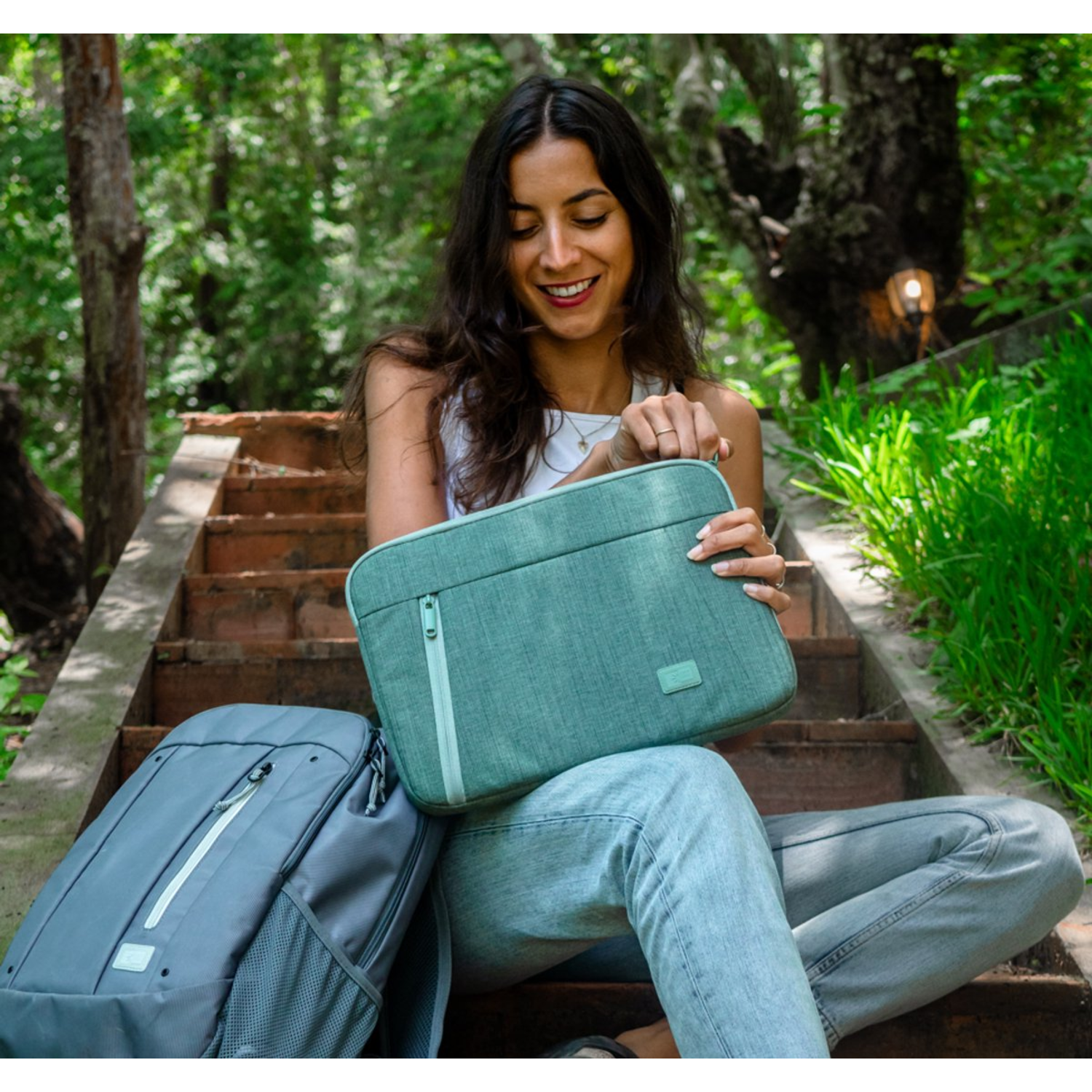 Femme sur escaliers tient étui ordinateur vert d'eau, sac à dos gris à côté. Fond forêt.