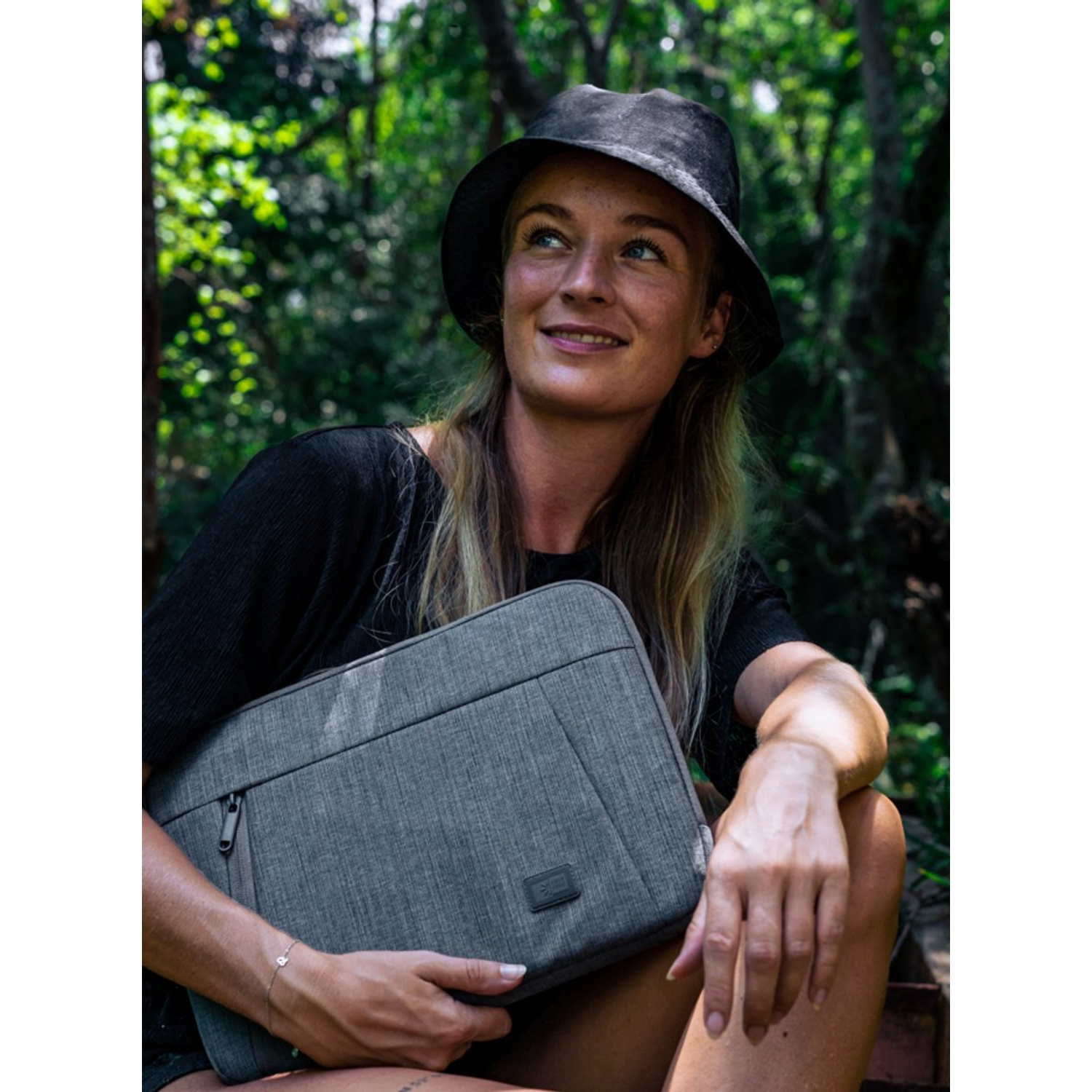 Une femme portant un chapeau noir tient une housse d'ordinateur portable grise, souriante, avec un fond de forêt. Elle est assise dehors.