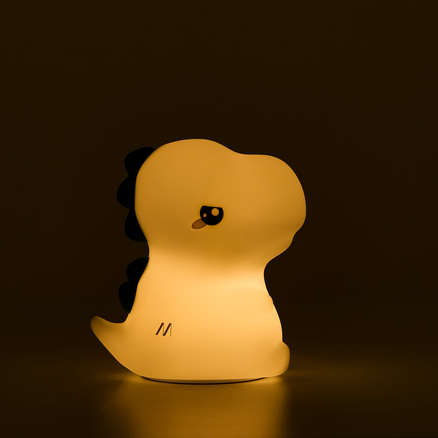 Una luce notturna a forma di dinosauro giallo incandescente con dettagli neri, un piccolo occhio e la lettera 'M'.
