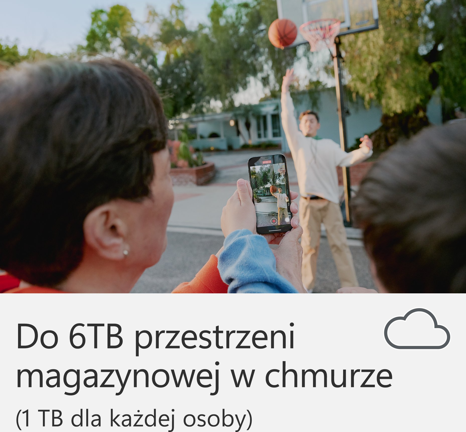 Osoba nagrywa mecz koszykówki na telefonie. Mężczyzna rzuca piłkę do kosza. Gra odbywa się na zewnątrz z drzewami.
