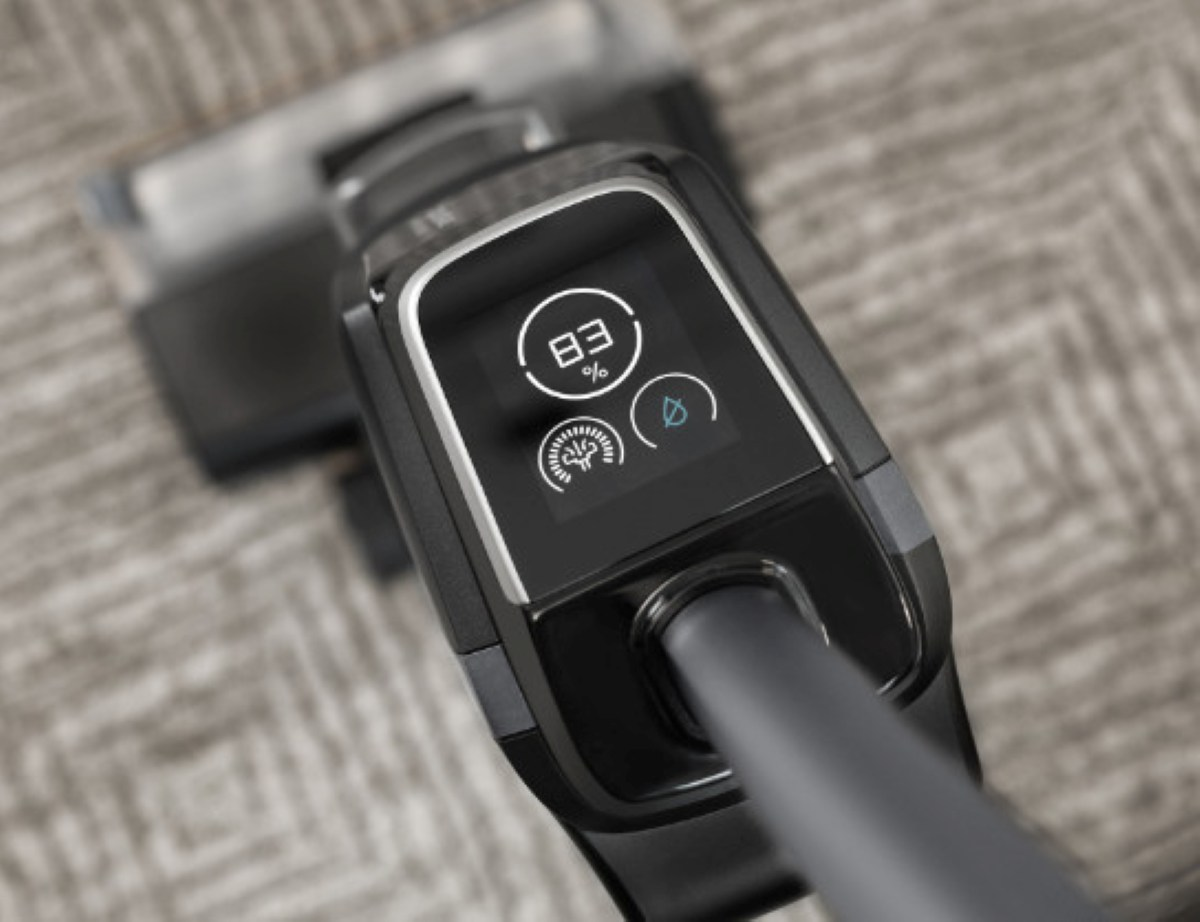 Zbliżenie czarnego uchwytu odkurzacza z ekranem wyświetlającym 83%, symbol wody i ikonę trybu czyszczenia. Tło to dywan.