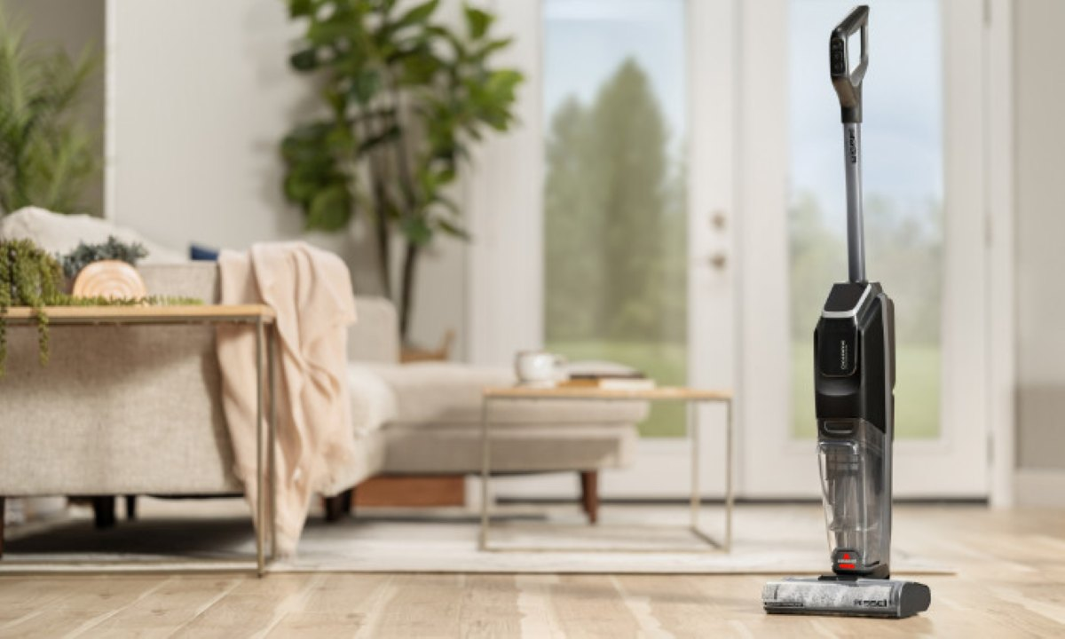 Czarno-szary odkurzacz stoi na drewnianej podłodze. W tle kanapa i rośliny w pobliżu okna.