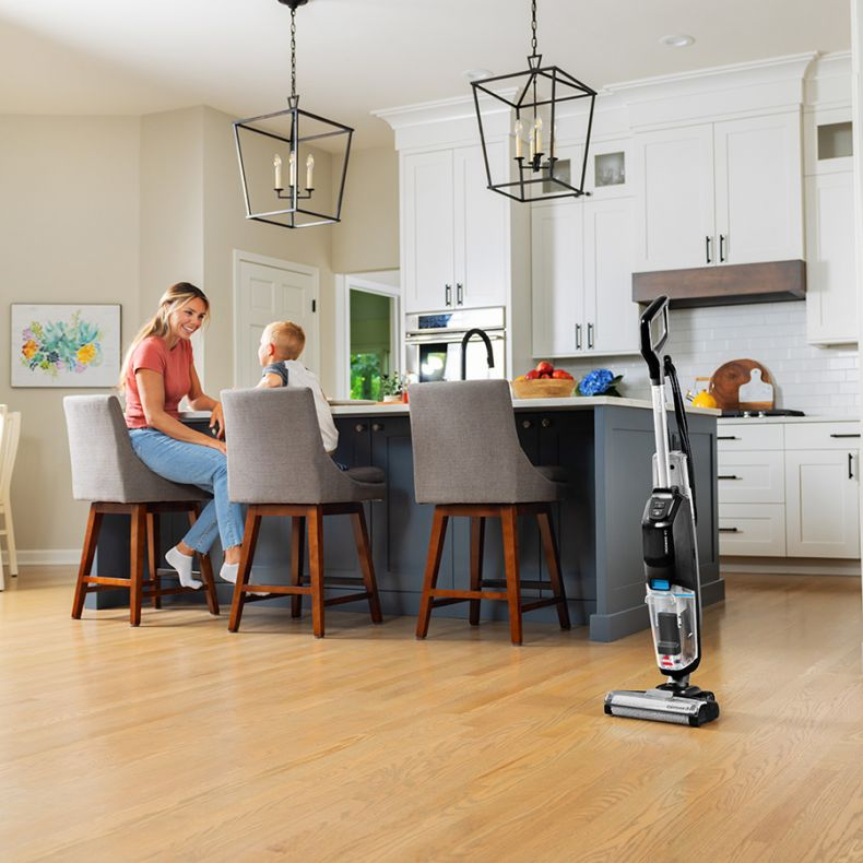 Kobieta i dziecko siedzą przy wyspie kuchennej. Odkurzacz stoi w pobliżu. Kuchnia ma białe szafki i drewniane podłogi.