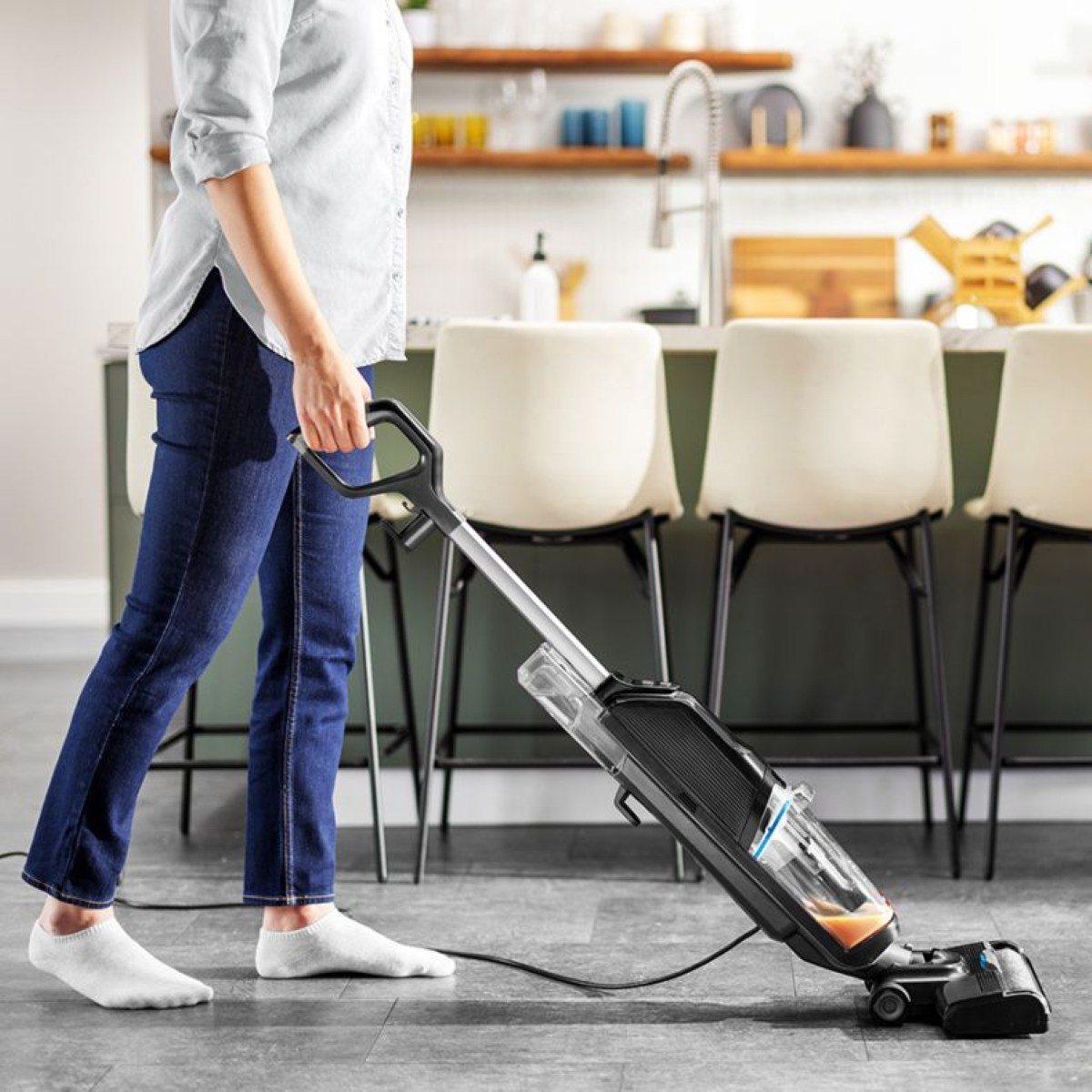 Osoba odkurza podłogę w kuchni. Ma na sobie dżinsy, białą koszulę i skarpetki. Widoczny jest odkurzacz i krzesła.