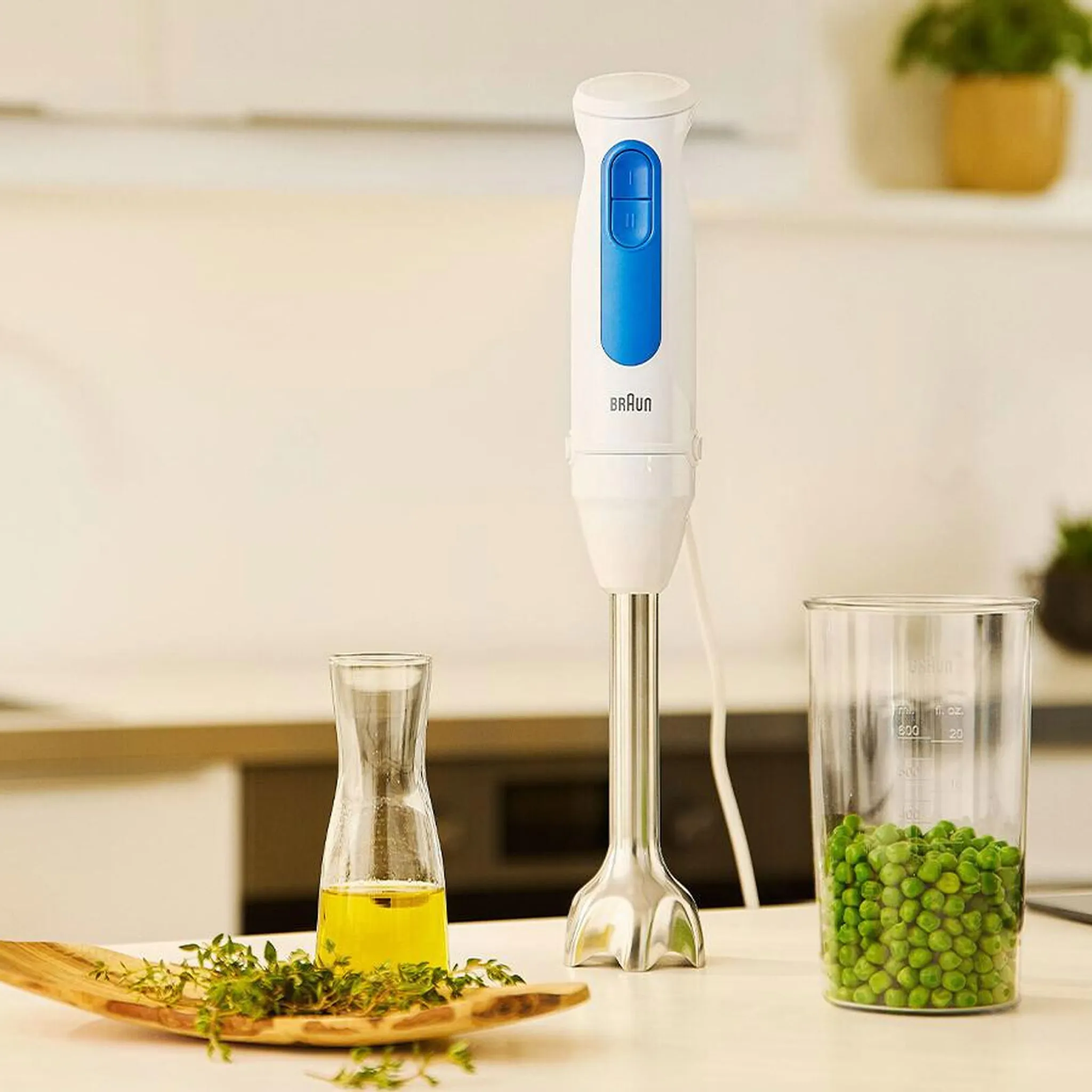 Biały i niebieski blender ręczny Braun stoi obok groszku w szklance. Na pierwszym planie oliwa z oliwek i zioła.