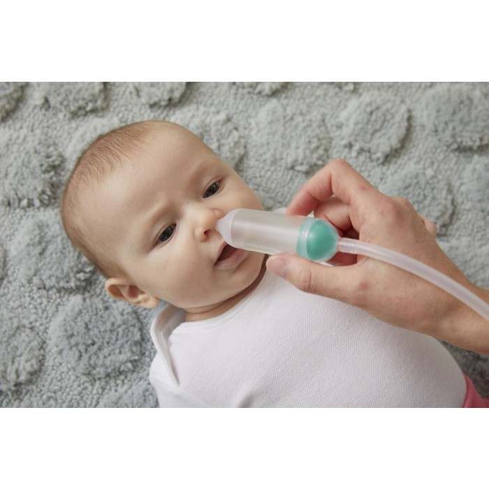 Dziecku odsysa się nos za pomocą urządzenia. Osoba trzyma urządzenie. Dziecko leży na szarej, wzorzystej kołdrze.